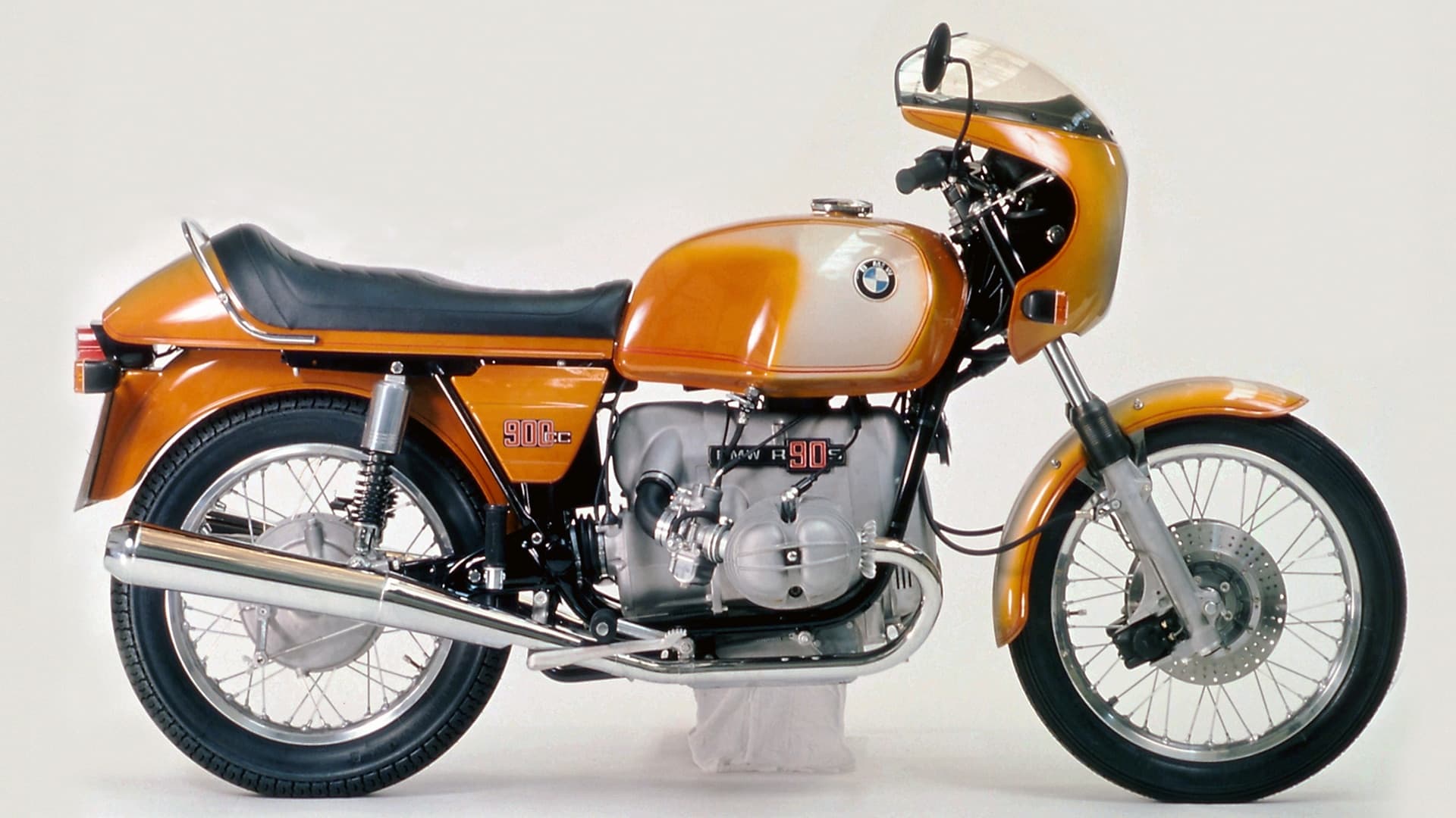 Side profile of a BMW R 90 S with orange and silver paint, black saddle and a small cockpit fairing.