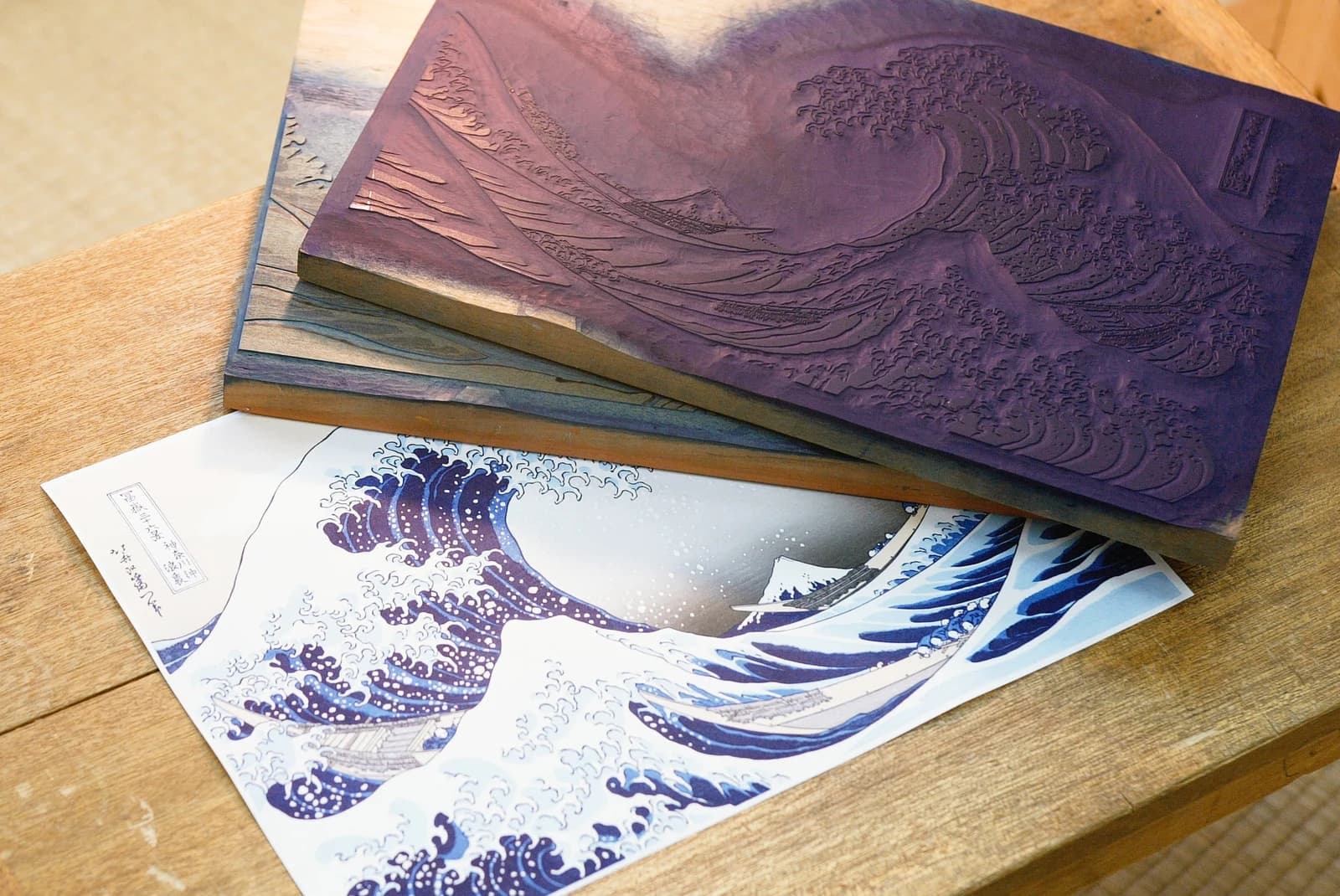 Carved woodblocks rest beside an Adachi reproduction of Hokusai’s Great Wave on a wooden table.