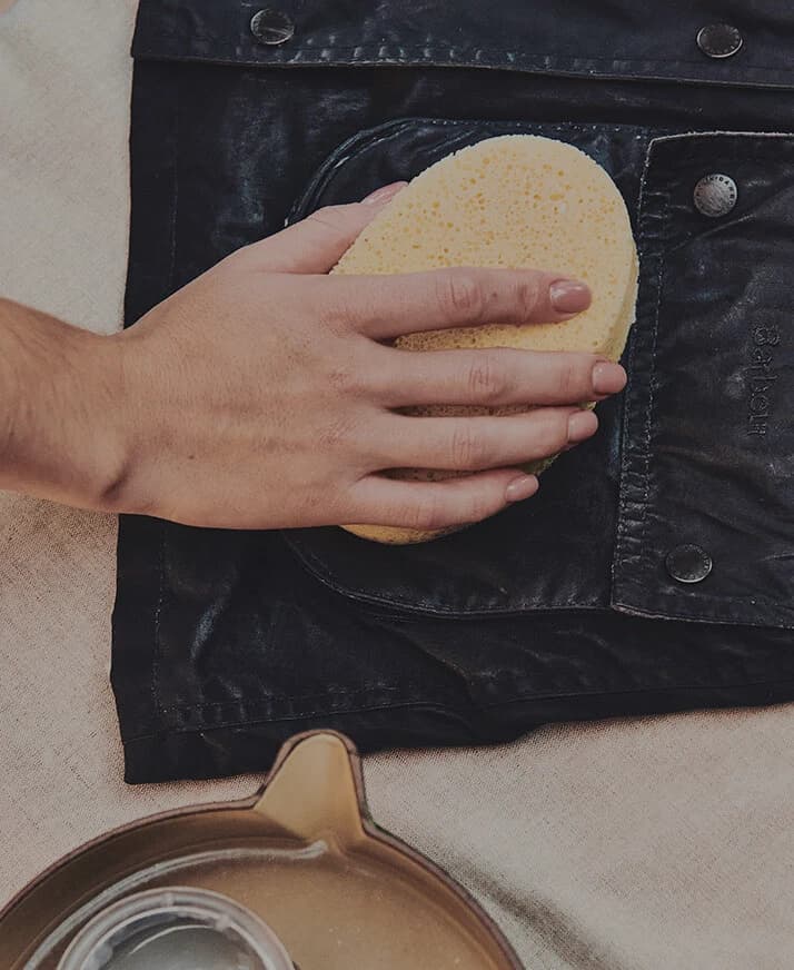 A hand applies wax with a sponge to the pocket area of a dark Barbour waxed jacket.