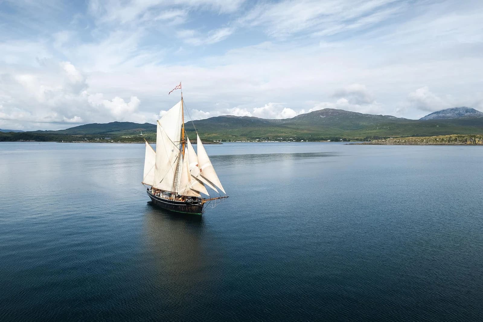 The traditional ketch Bessie Ellen under sail on calm blue water with Hebridean hills beyond.