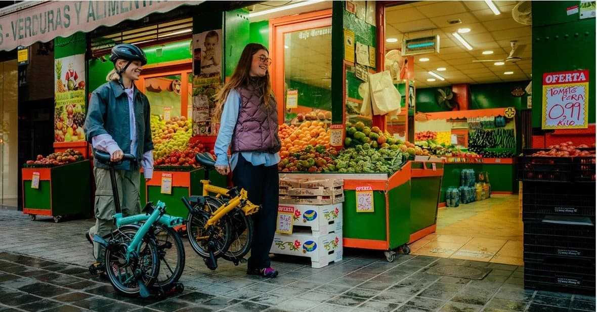 Two people carry folded turquoise and yellow Brompton C Line bicycles past a greengrocer.