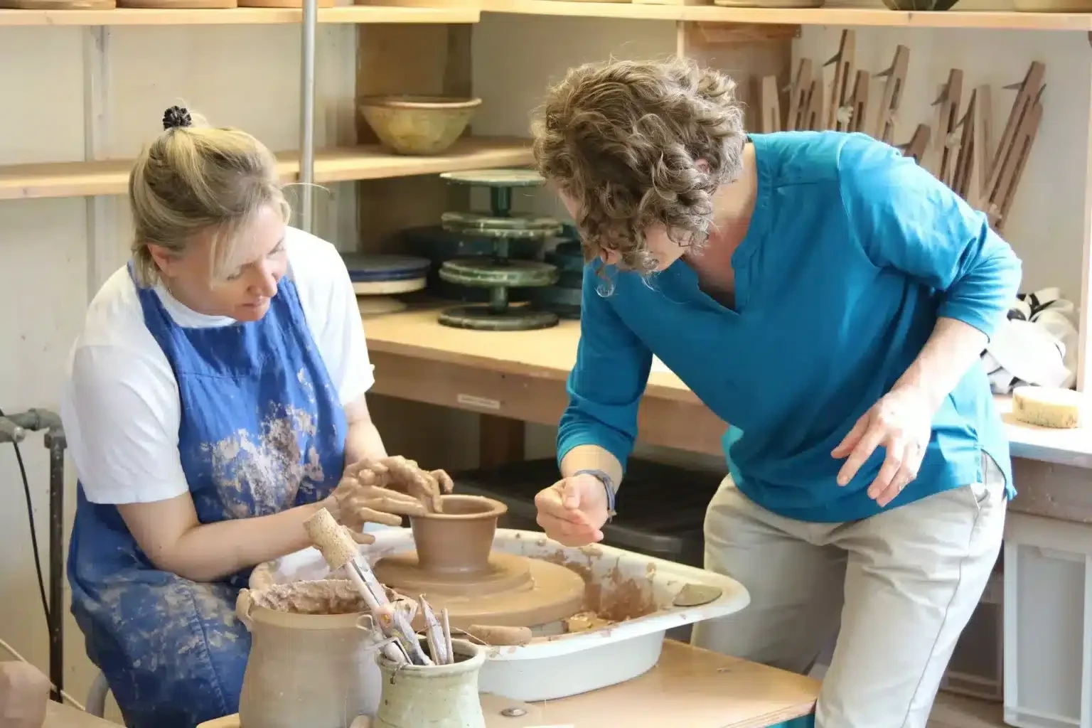 A tutor helps an adult potter shape a clay cylinder on an electric wheel at the Leach Pottery.