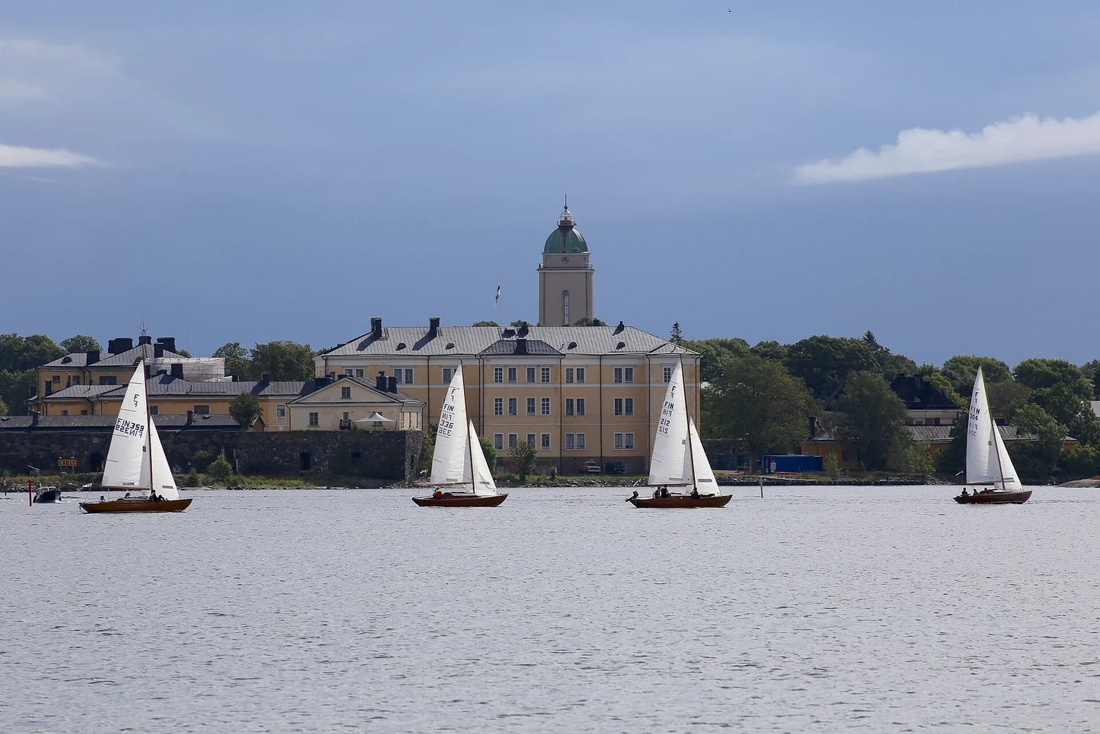Nordic Folkboats sail through Helsinki waters with a coastal fortress beyond their white sails.