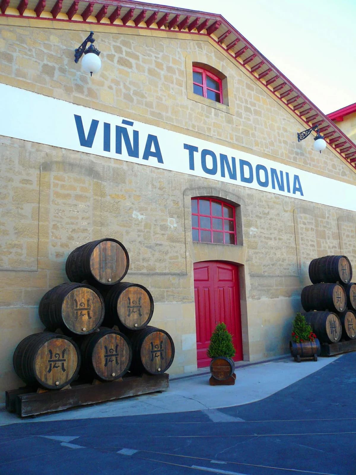 Oak barrels flank a red door beneath the Viña Tondonia name on a pale stone winery building in Haro.