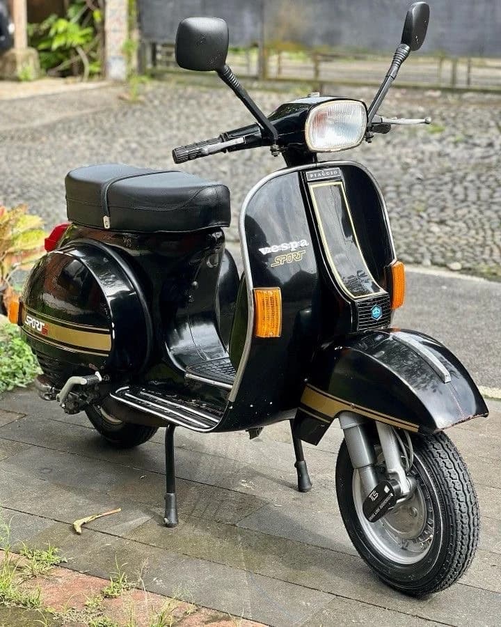 A black Vespa PX Sport with gold detailing stands on paving, photographed from the front quarter.