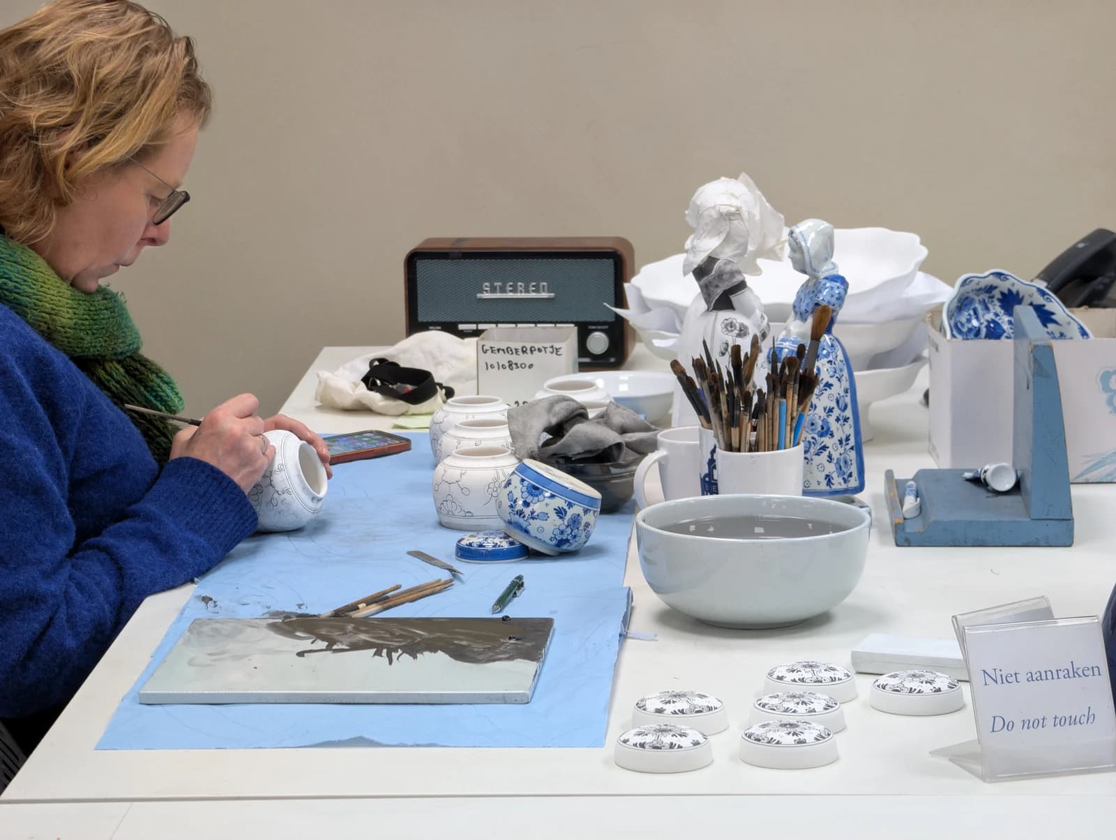 A painter at Royal Delft works on a small ceramic jar beside brushes, dark pigment and blue finished pieces.