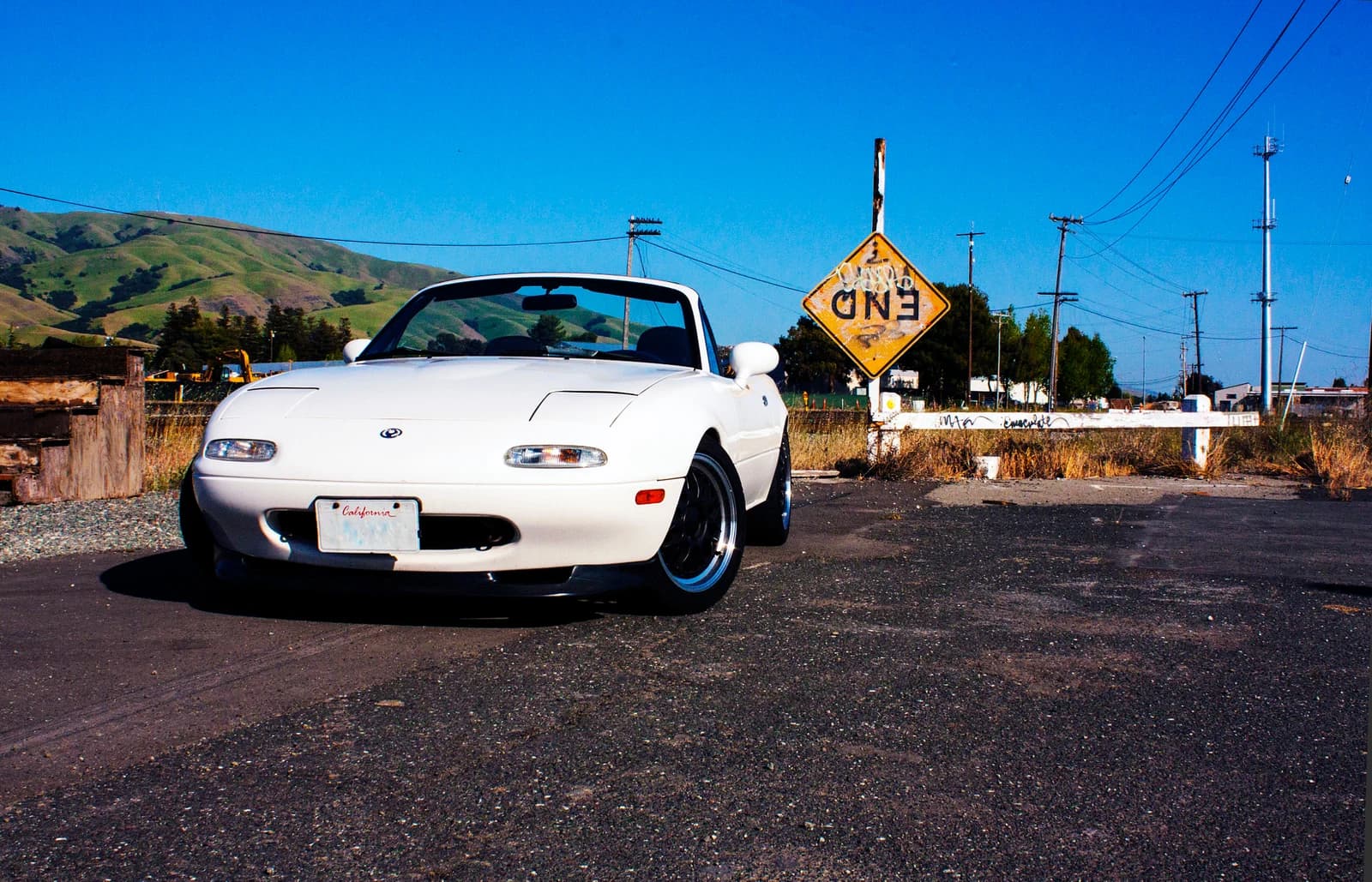 A white first-generation Mazda MX-5 roadster with its roof down on a quiet road beneath a deep blue sky.