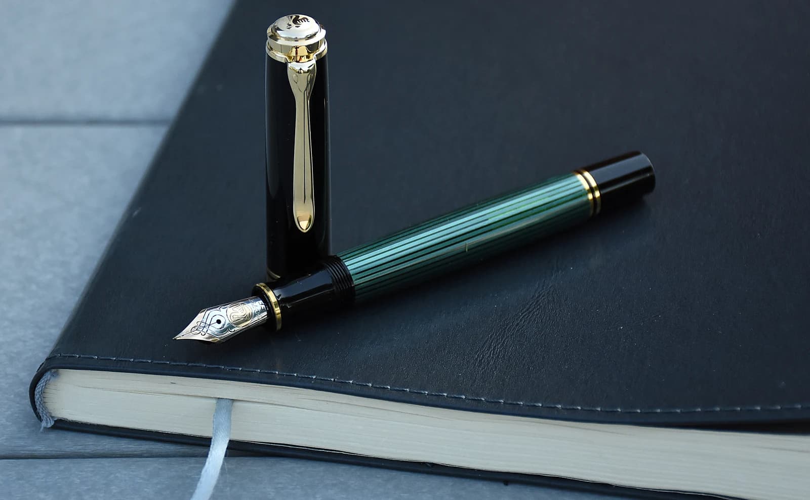 A green-striped Pelikan M800 fountain pen with its nib exposed and cap upright on a dark notebook.