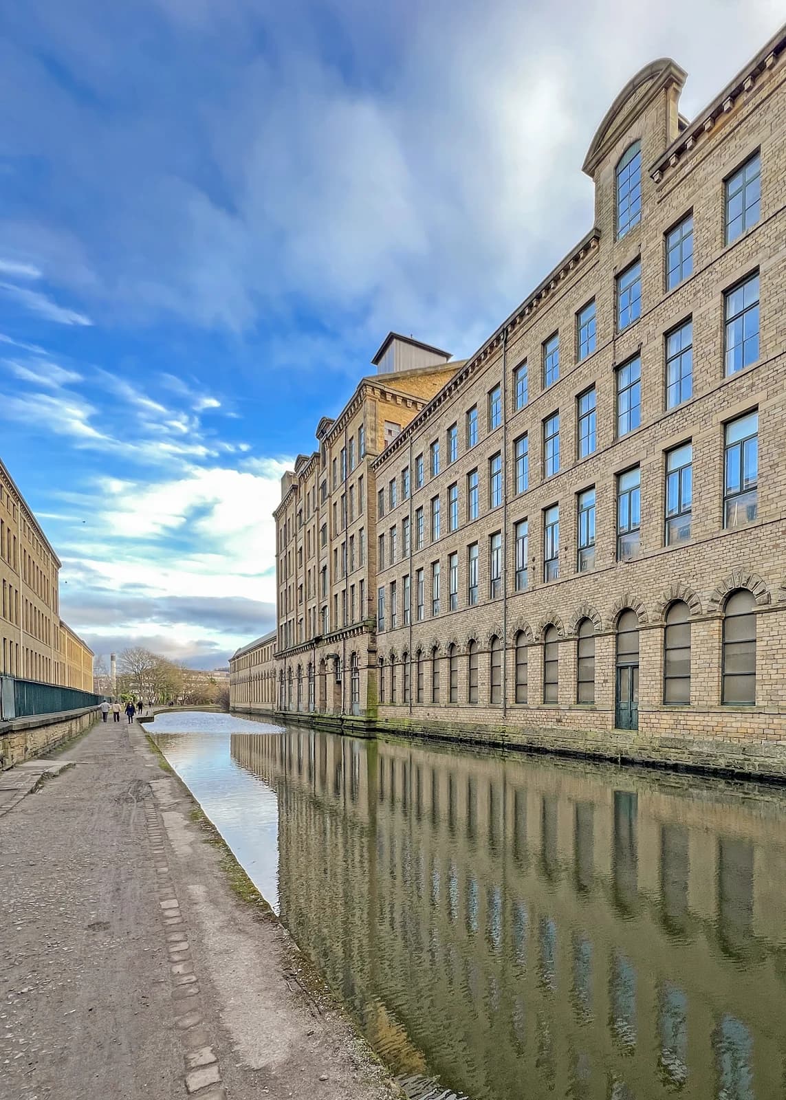The long stone facade of Salts Mill follows the Leeds and Liverpool Canal beneath a blue-grey sky.