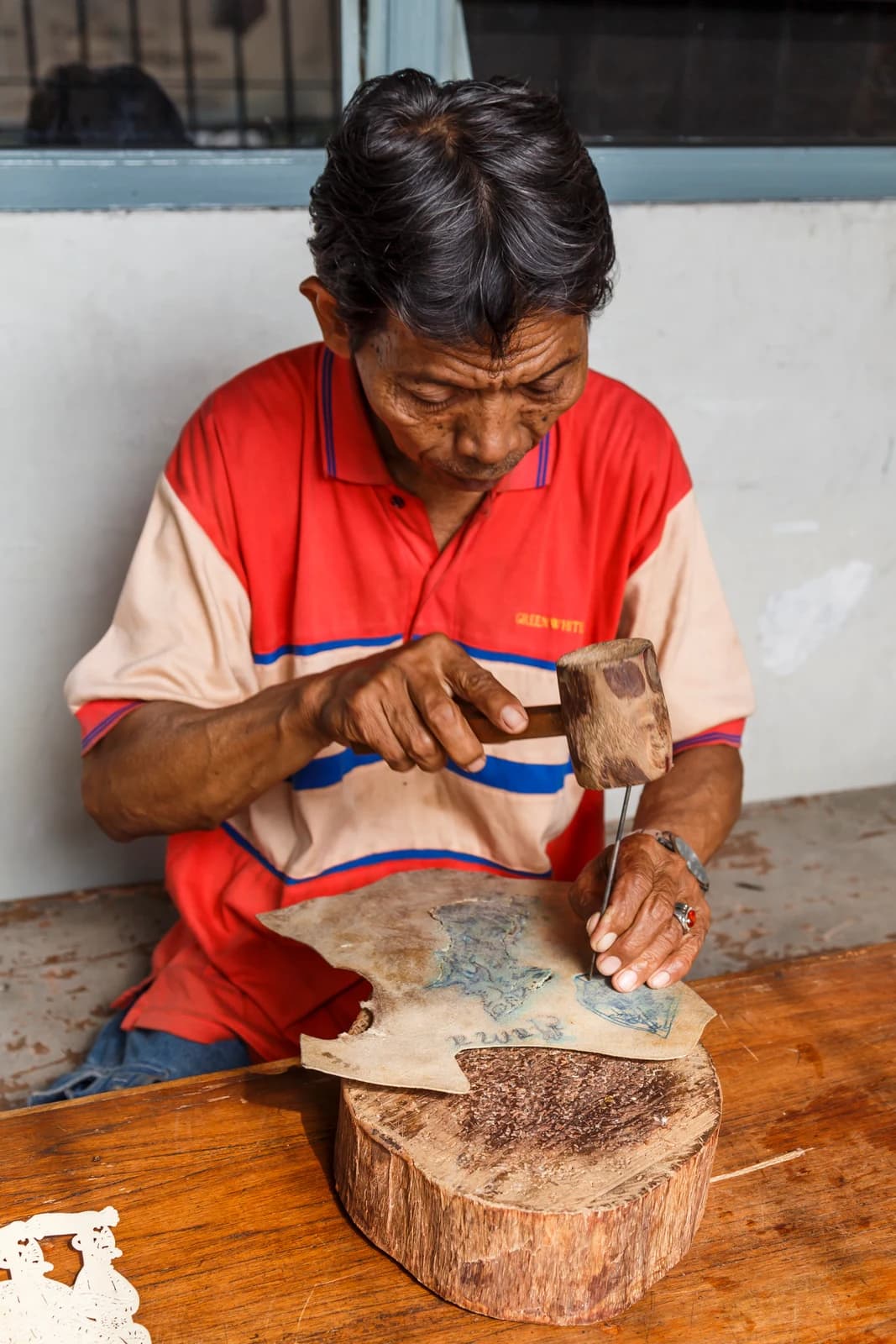 A wayang kulit craftsperson in Jakarta uses a mallet and fine tool to work leather on a wooden block.