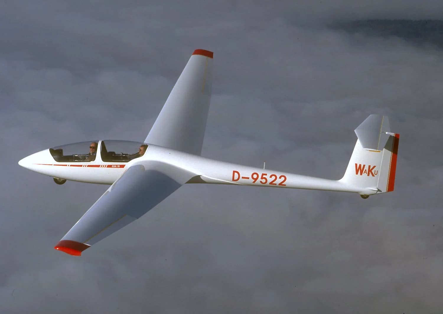 A white Schleicher ASK 21 two-seat sailplane banks above a clouded landscape with both seats visible beneath its canopy.