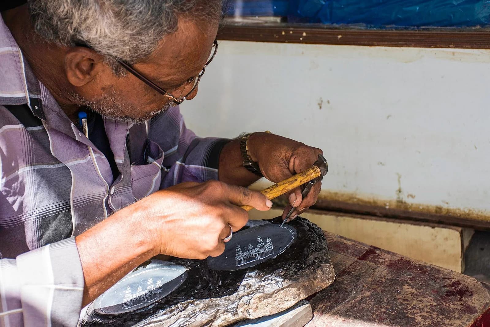 A craftsperson leans close to a dark Bidriware plate while applying fine inlaid metal detail by hand.