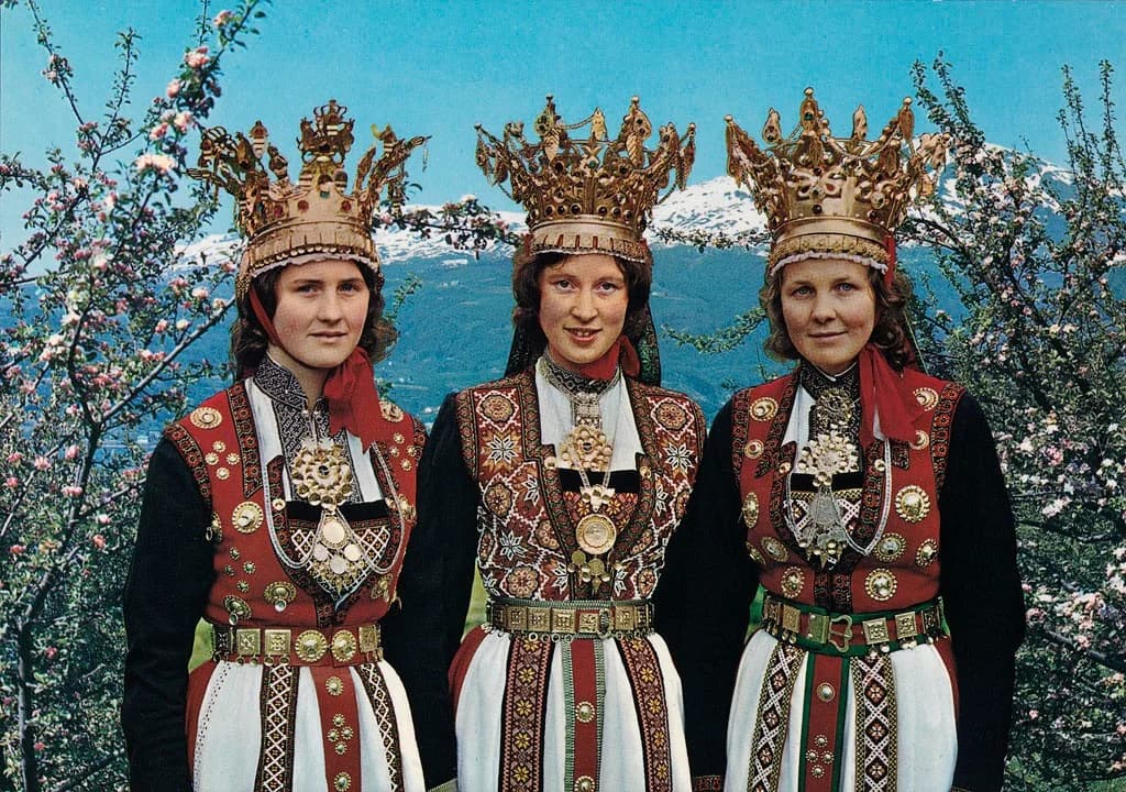 Three women outdoors in historical Hardanger bridal costumes with embroidered dress and elaborate bridal crowns.
