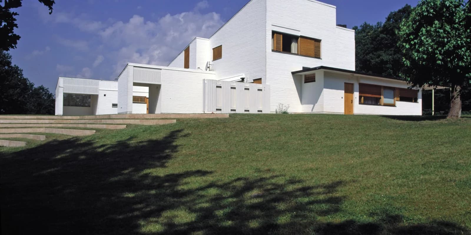 The white brick volumes and timber shutters of Maison Louis Carré step across a green slope beneath mature trees.
