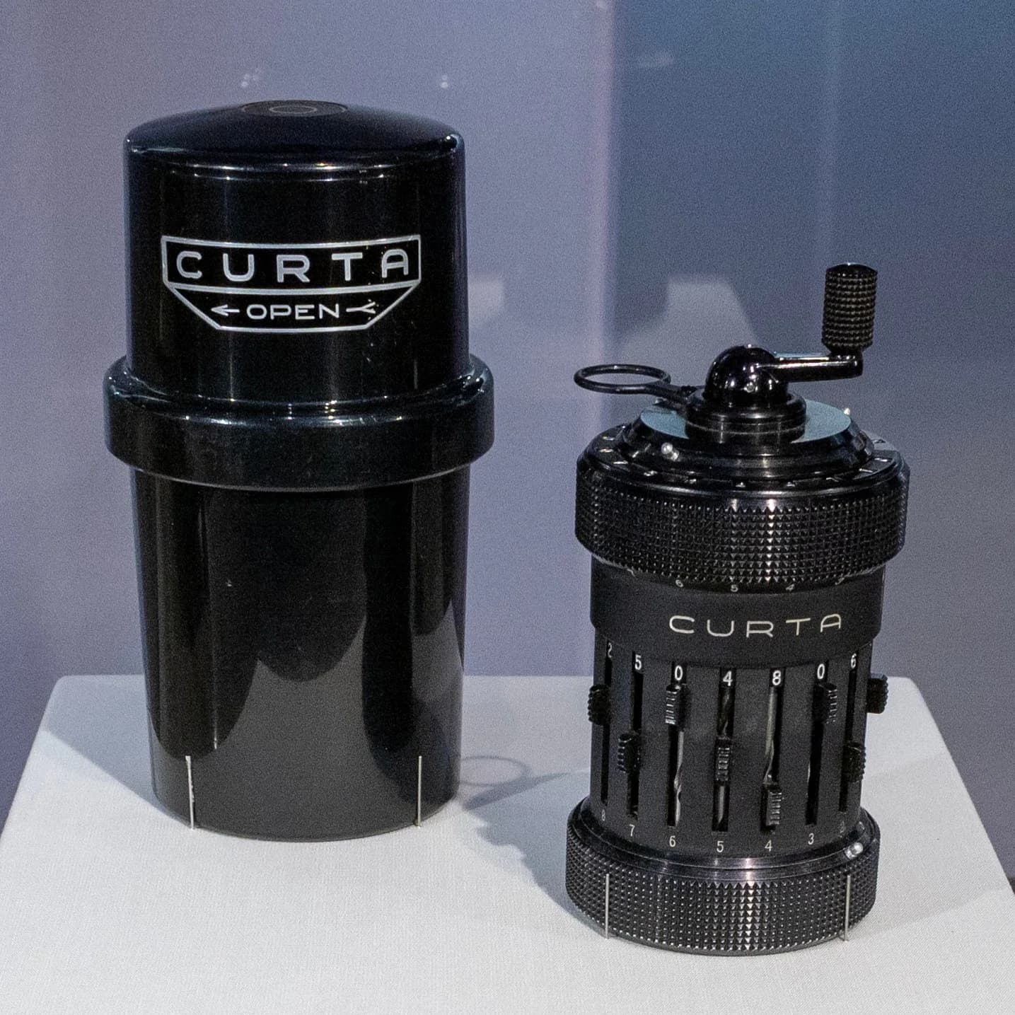 A black cylindrical Curta mechanical calculator and matching case on a plain museum display surface.