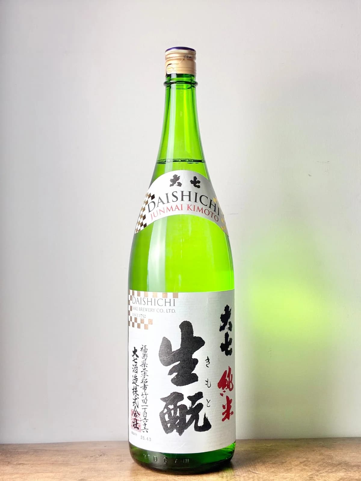 A green bottle labelled Daishichi Junmai Kimoto stands upright against a neutral wall on a wooden surface.