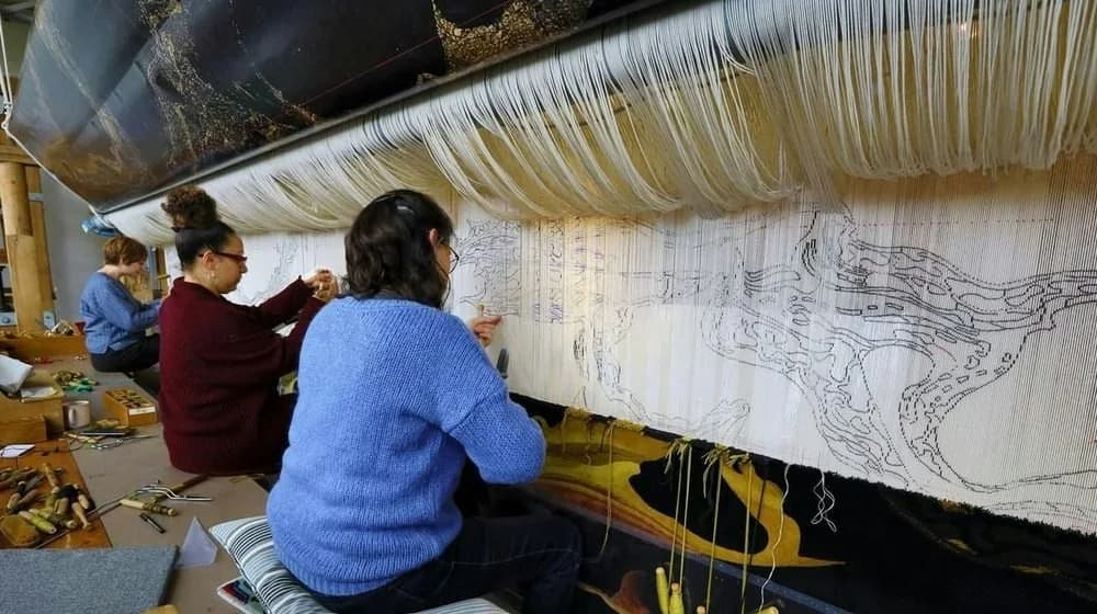 Three tapestry weavers work in a bright Paris atelier, with drawn lines visible through a broad field of pale warp threads.