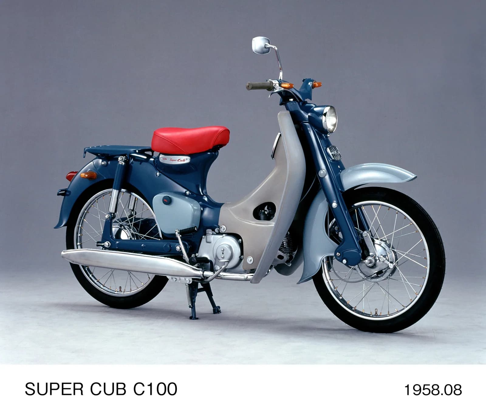 A blue and pale grey 1958 Honda Super Cub C100 in three-quarter profile, showing its low frame, leg shields and red seat.