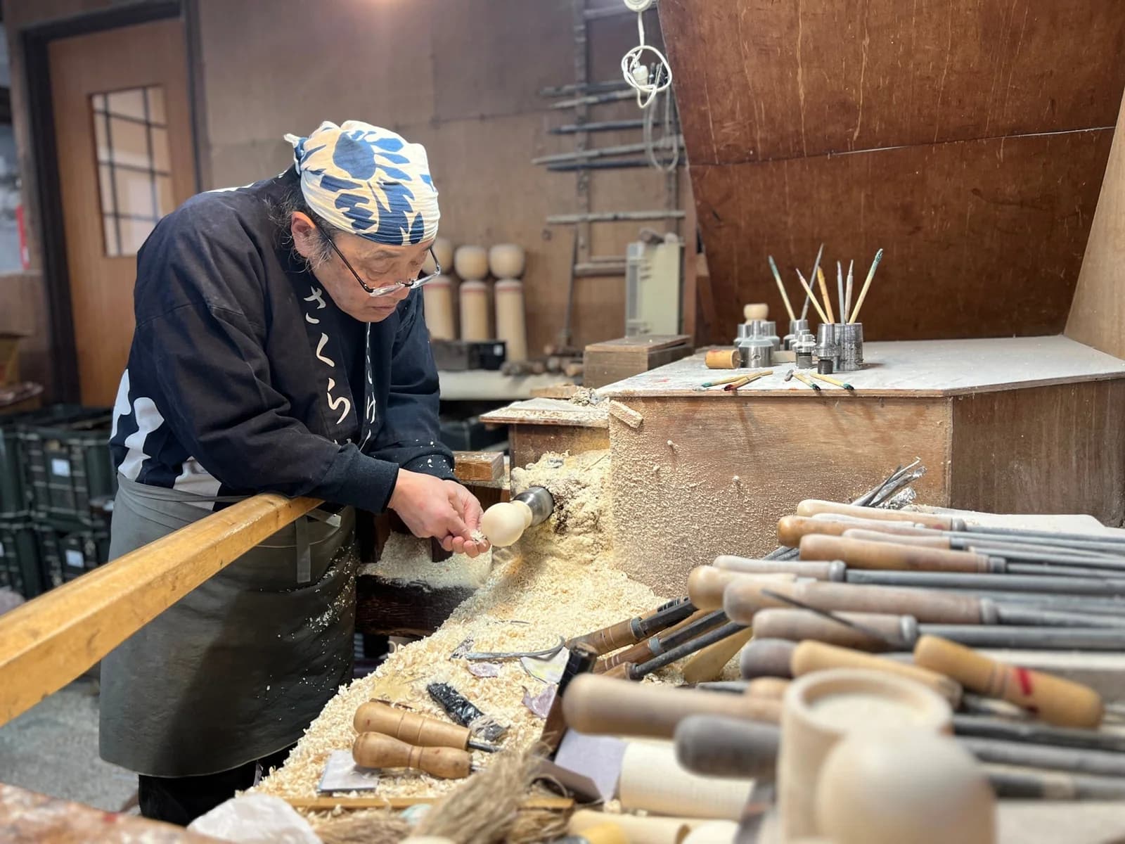 Akihiro Sakurai turns a wooden kokeshi form at his Naruko workshop, surrounded by tools and pale wood shavings.