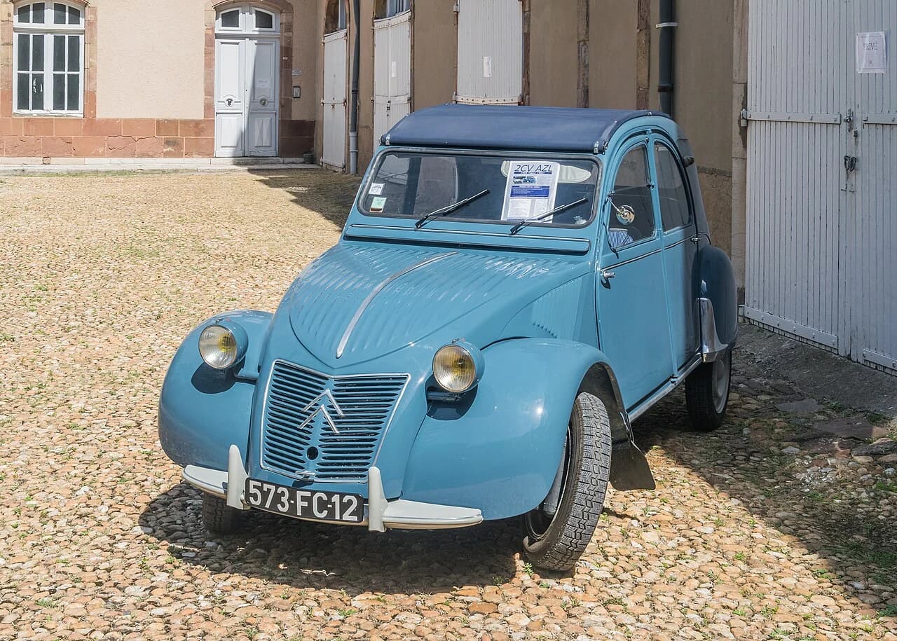 A blue Citroën 2CV with a folding fabric roof stands on gravel in a three-quarter front view.
