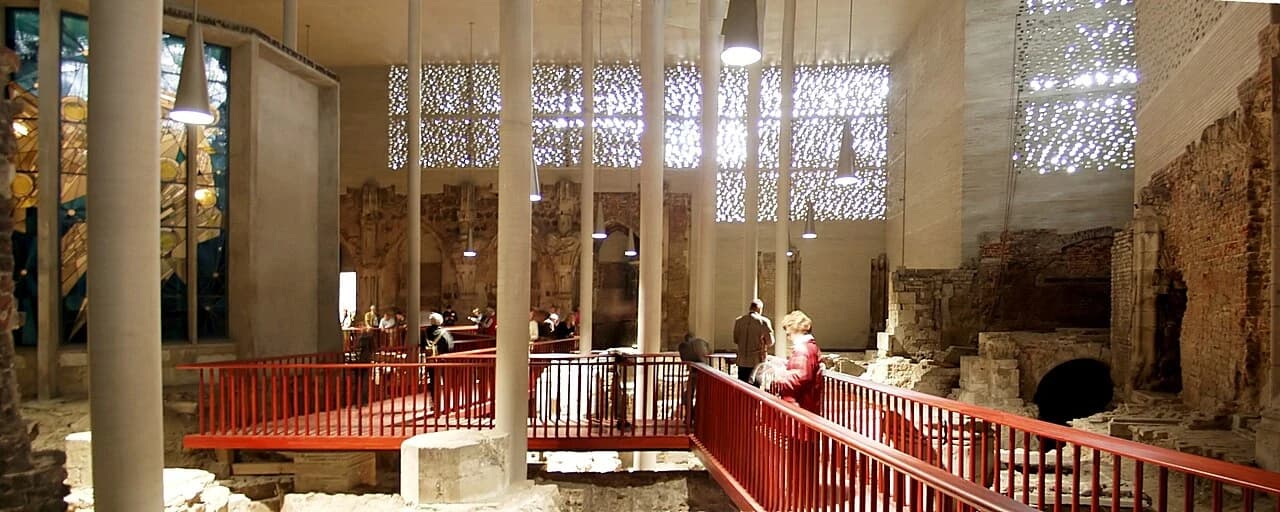 A panoramic view of the archaeological remains inside Kolumba’s tall contemporary hall, with a walkway passing above old stone walls.