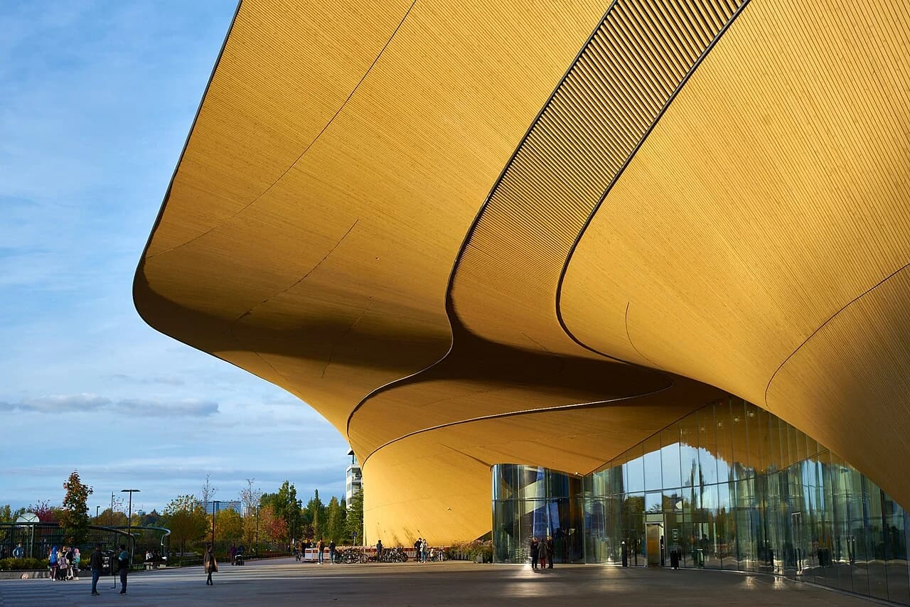 The curving timber facade of Helsinki’s Oodi library sweeps over a paved public space beneath a bright blue sky.