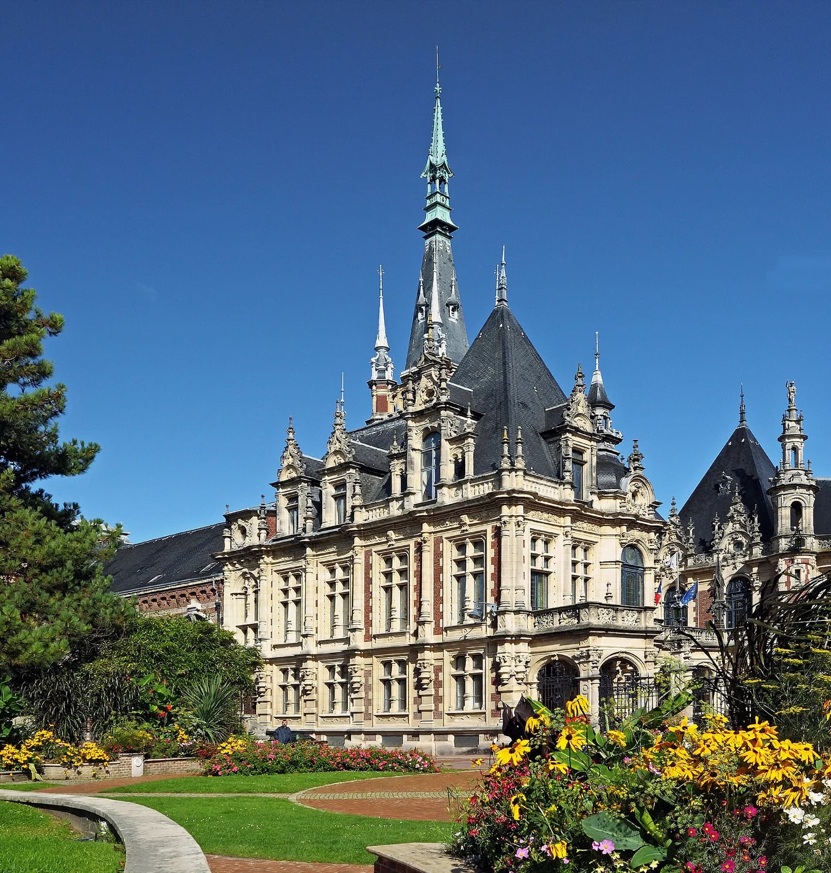 The ornate Palais Bénédictine at Fécamp rises above flowering gardens beneath a vivid blue sky, with its central spire visible.