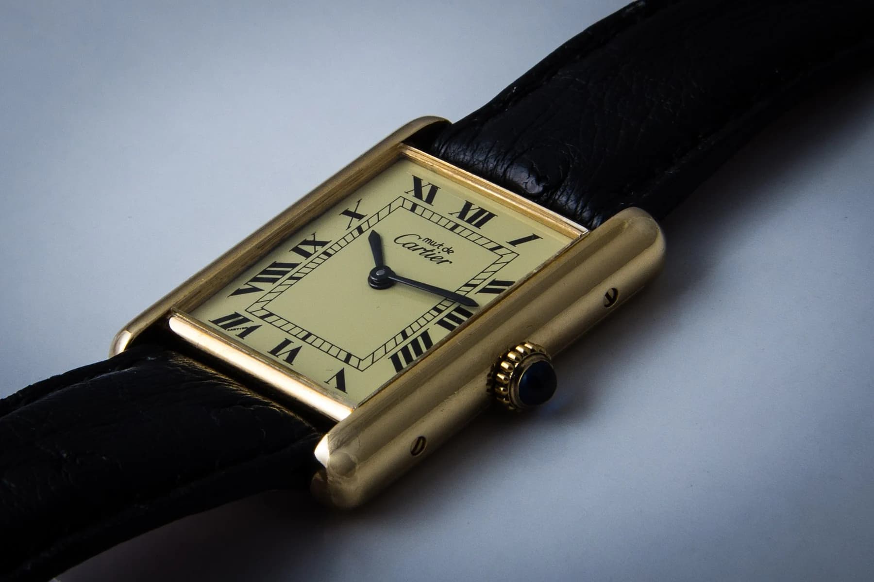 A gold-coloured Cartier Tank with a cream Roman-numeral dial and black leather strap lies diagonally on a pale surface.