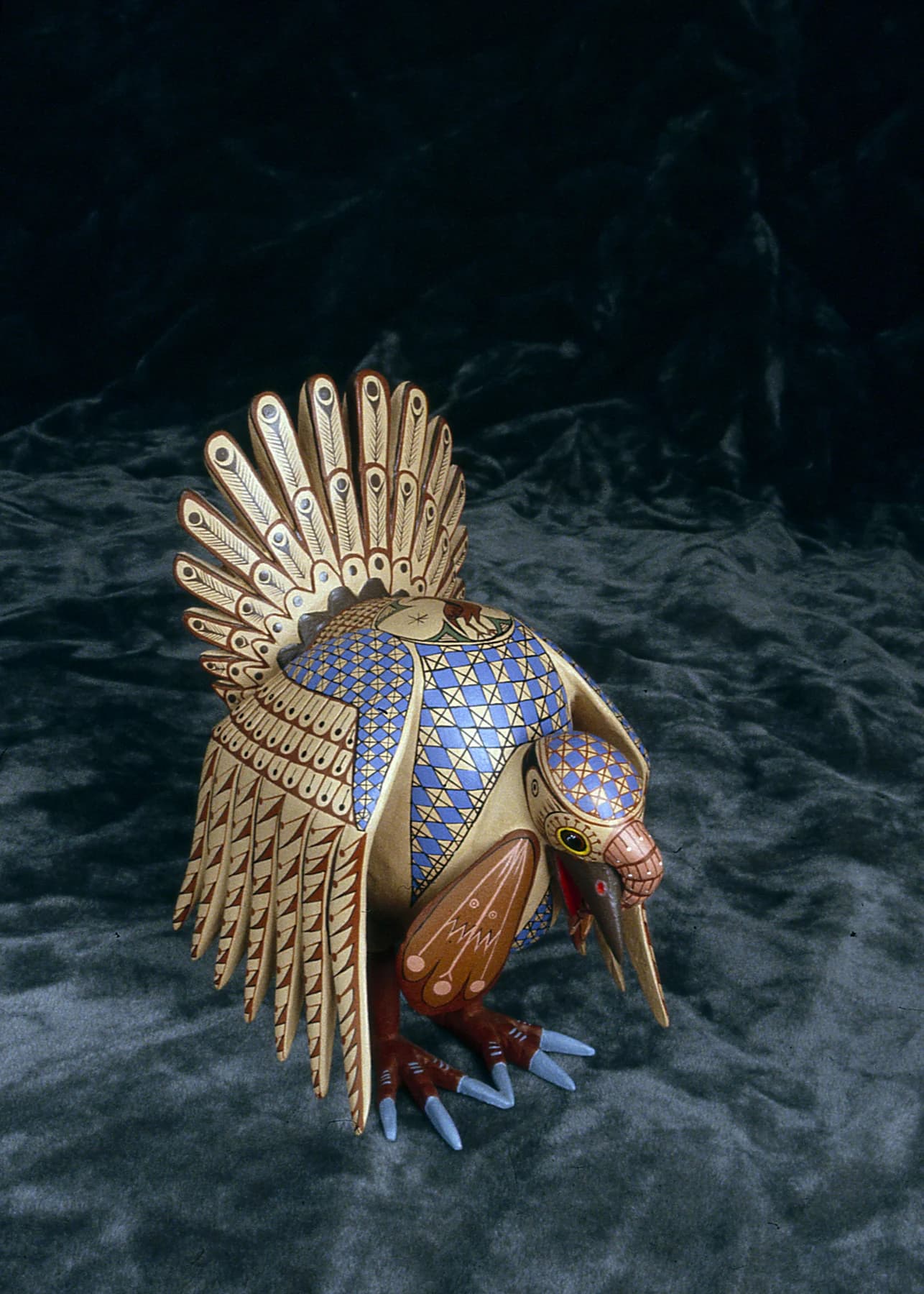 A richly patterned carved turkey by Jacobo Ángeles Ojeda stands against a dark blue background, with a fan-shaped gold and white tail.