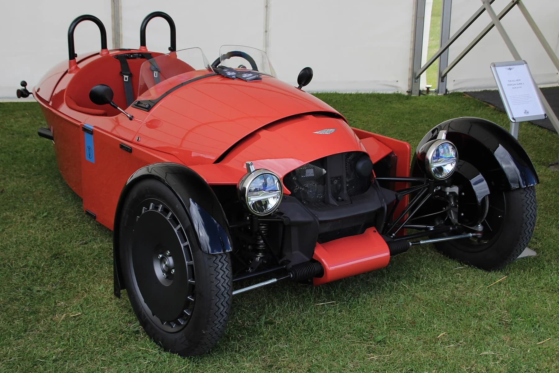 A red Morgan Super 3 with its two exposed front wheels and open two-seat cockpit stands on grass at a vehicle event.