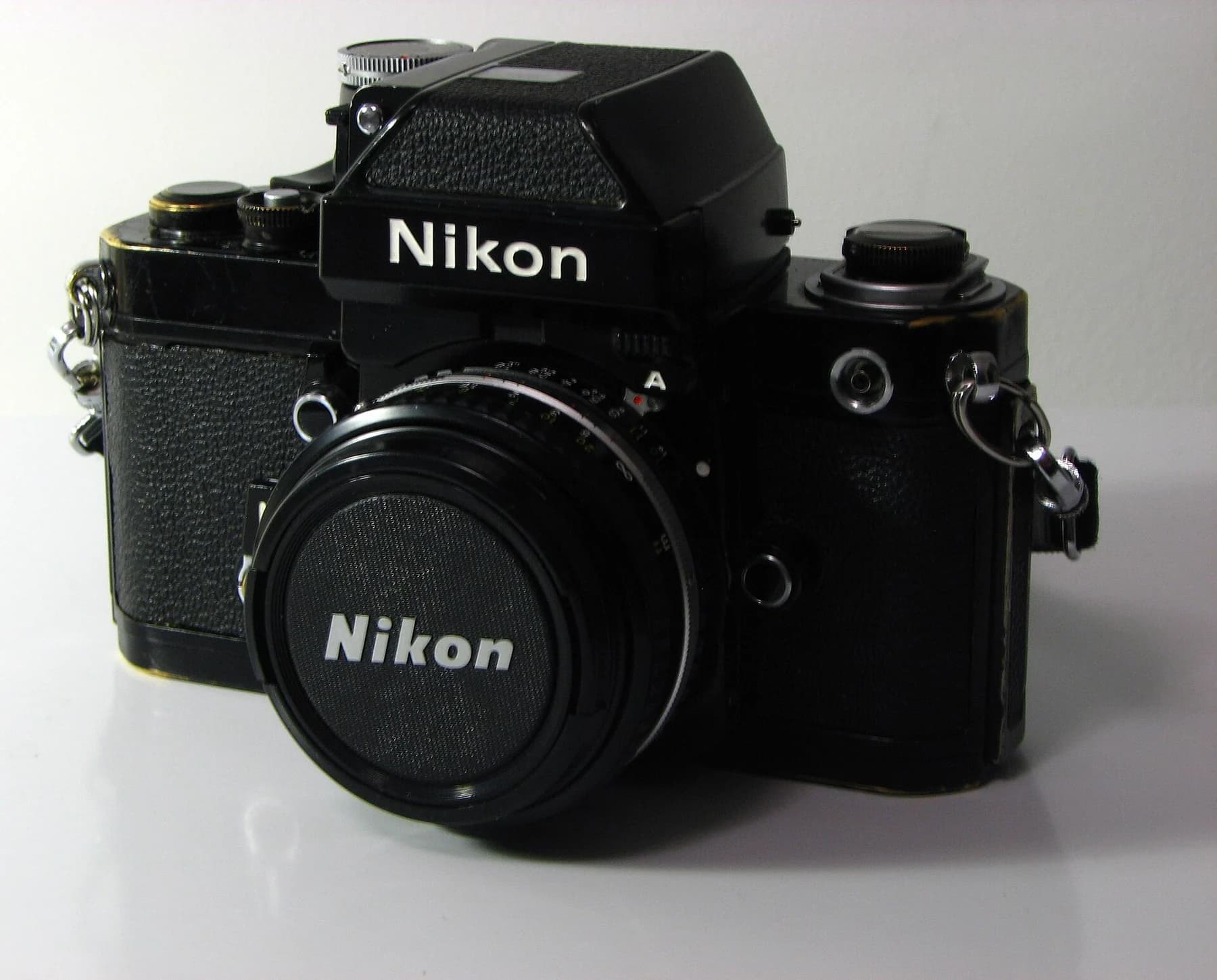A black Nikon F2 camera with a removable metering finder and capped lens, photographed in a three-quarter view on a pale surface.