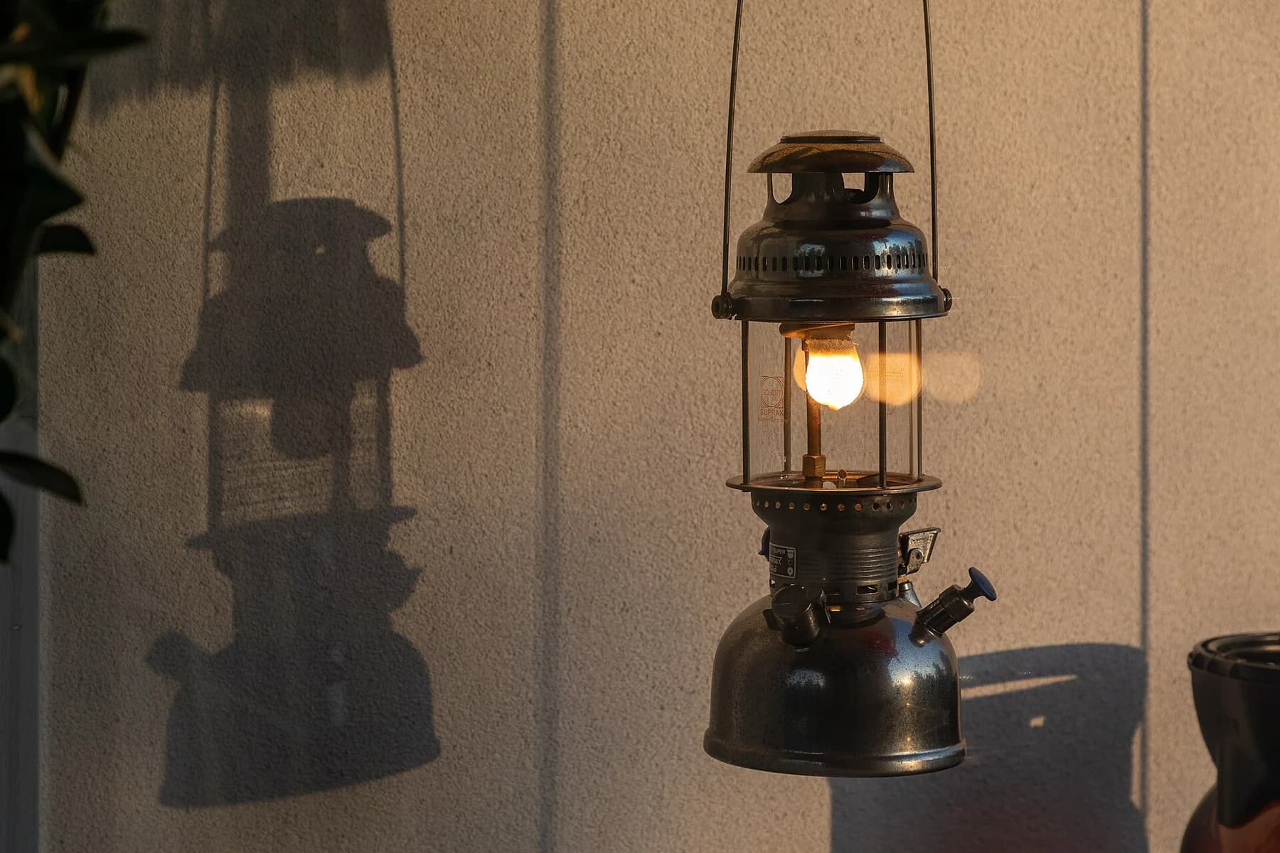 A patinated Petromax pressure lantern glows beside a textured wall at sunset, casting a long shadow of its shape.