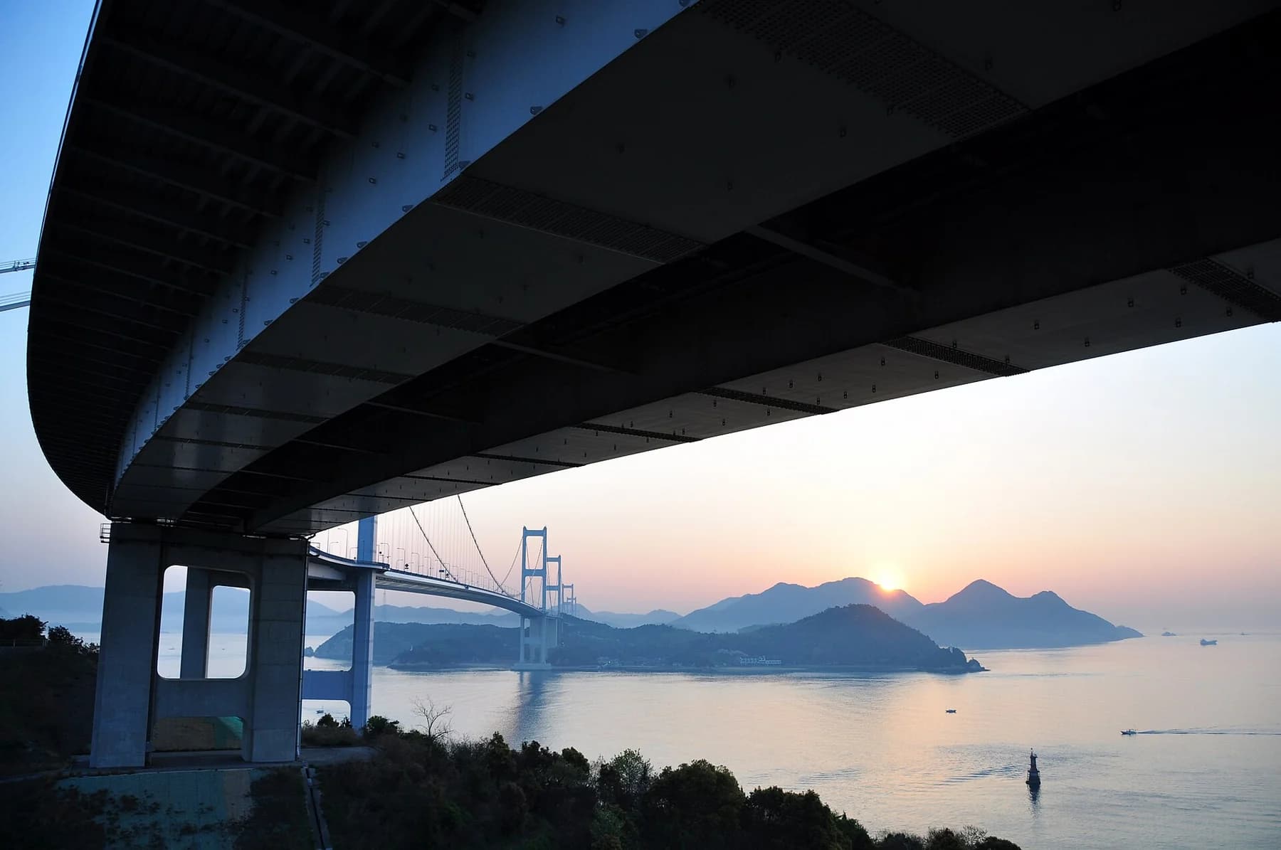 The Kurushima-Kaikyo bridges extend across the Seto Inland Sea at sunset, seen beneath one span with islands beyond.