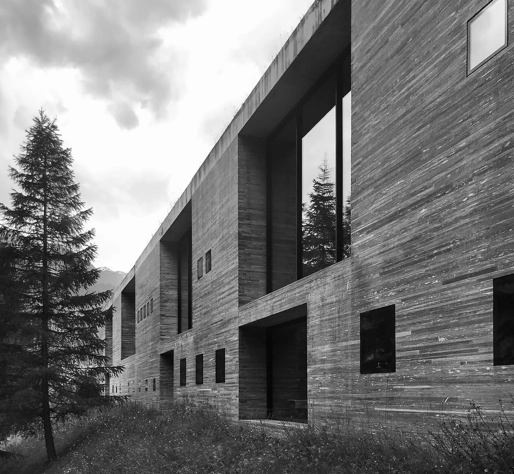 A black-and-white exterior view of Therme Vals shows its layered stone facade, tall openings and a tree under cloudy sky.