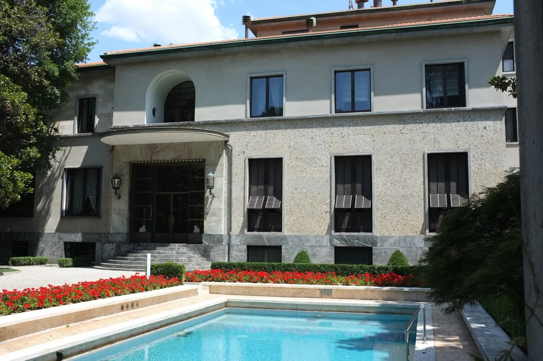 Villa Necchi Campiglio’s pale facade and veranda face a blue swimming pool framed by a garden and red flowers in Milan.