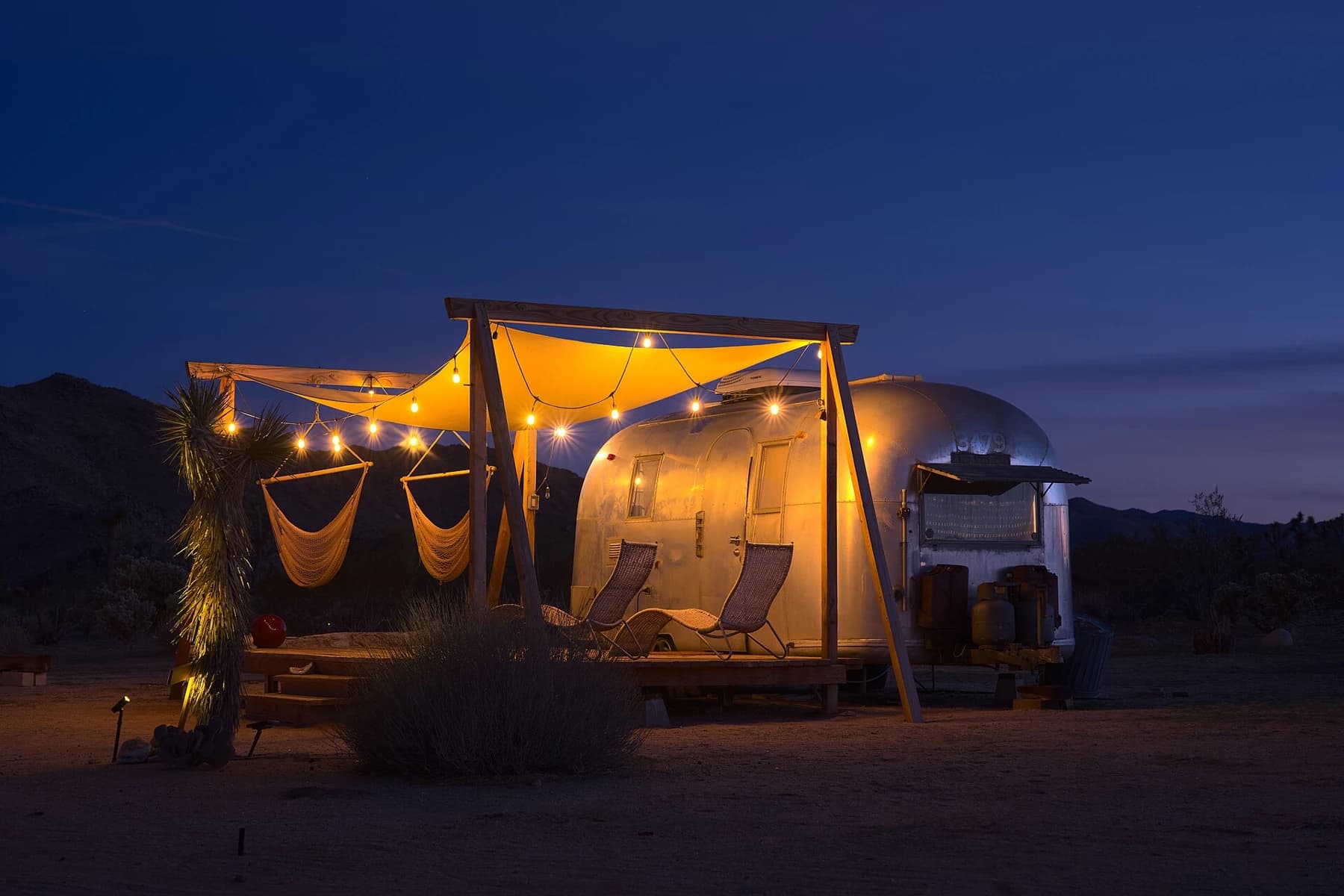A silver Airstream Safari trailer glows beneath a fabric canopy at dusk in Joshua Tree, with deep blue sky above the desert setting.