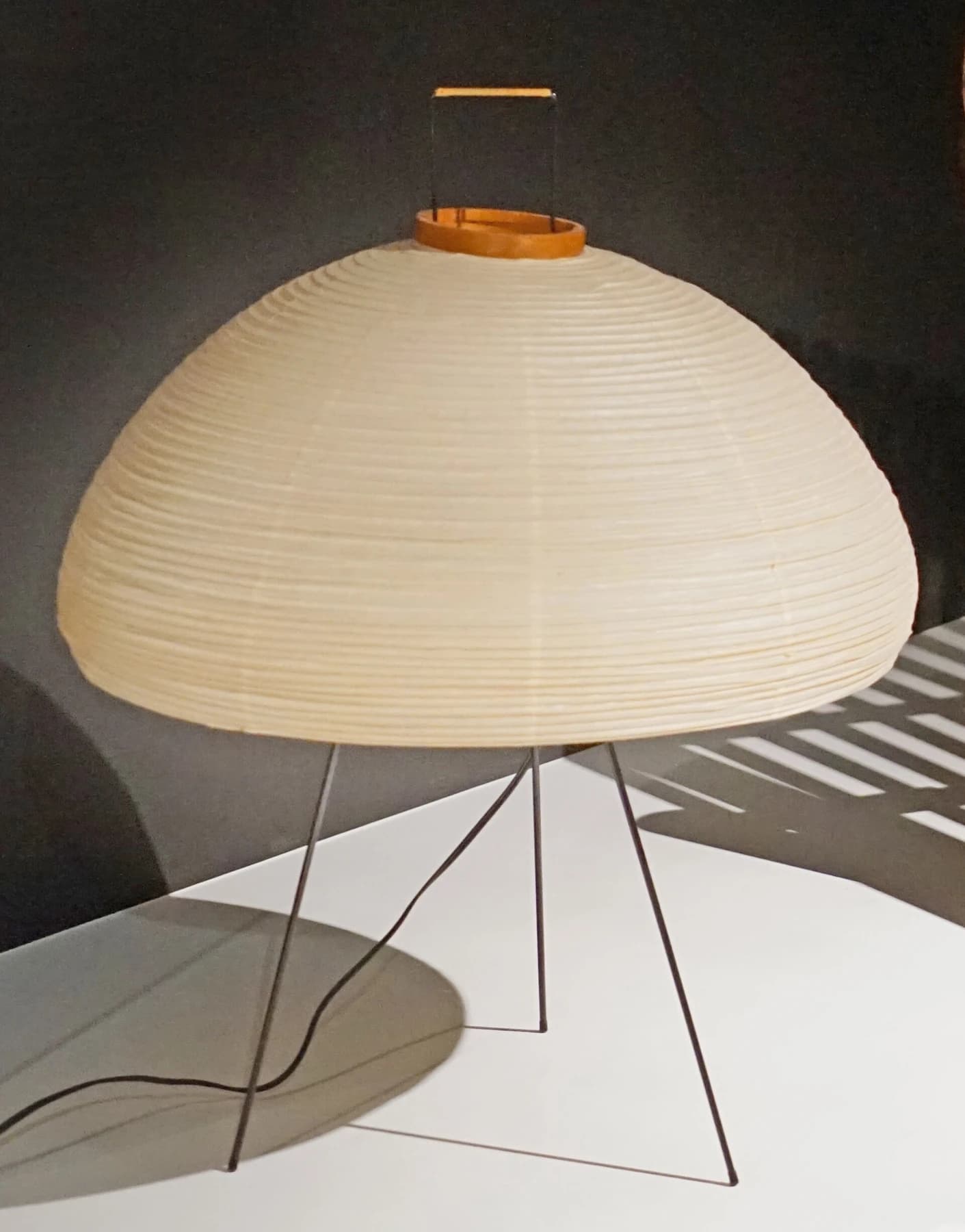 An Akari 10A lamp by Isamu Noguchi, with a rounded ribbed paper shade and slender metal legs, displayed in a Paris museum.