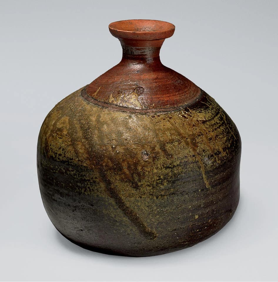 A rounded historical Bizen sake bottle with a narrow reddish neck and mottled brown, amber and dark surfaces, isolated on a pale background.