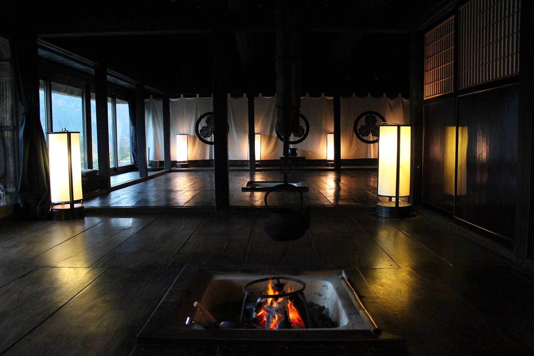 Chiiori’s dark timber interior with a recessed irori hearth, glowing standing lights and daylight entering along one side of the room.