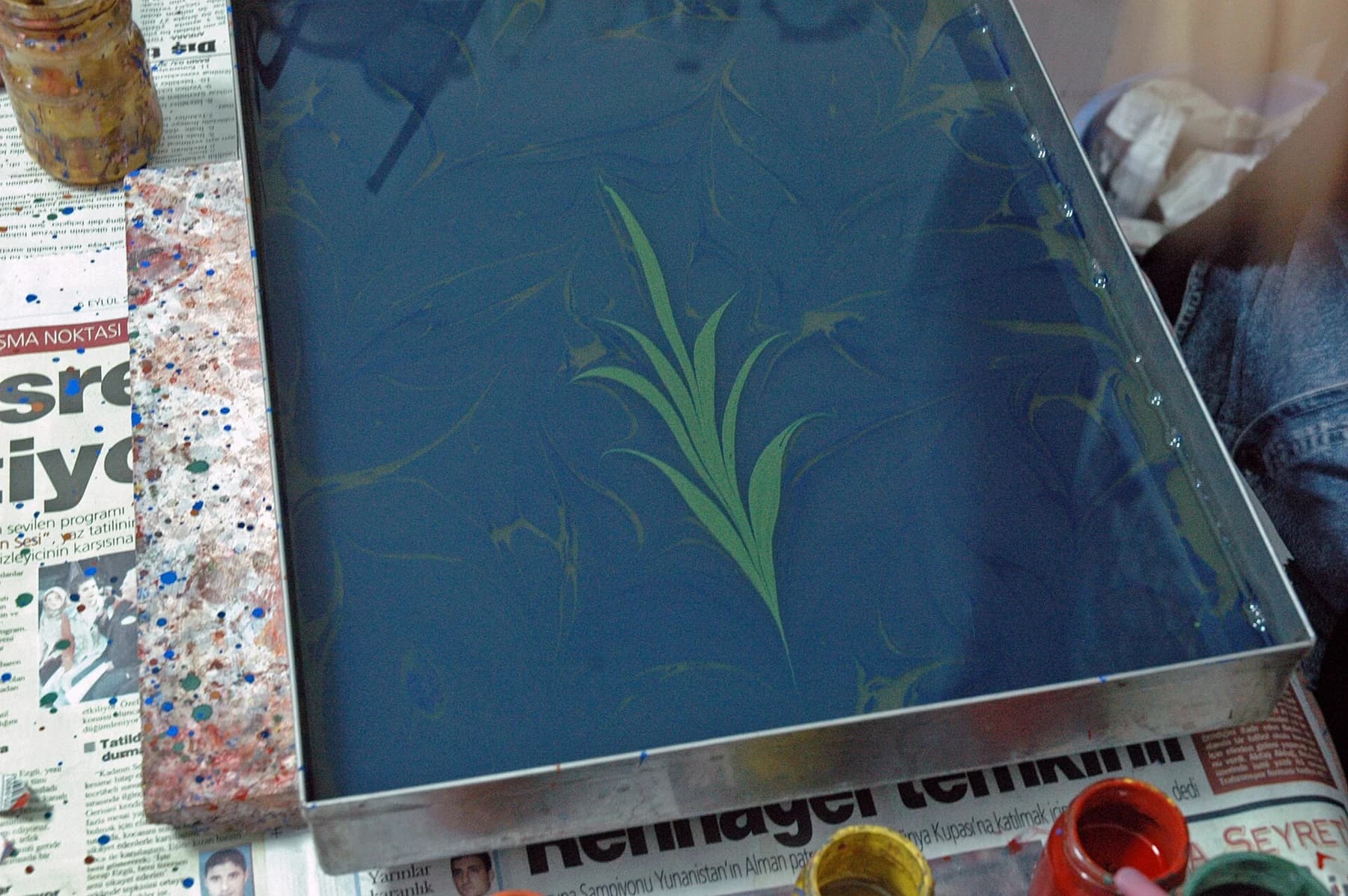 A green plant motif floats on a blue marbling bath inside a rectangular tray, with a hand at the edge and working materials around it.