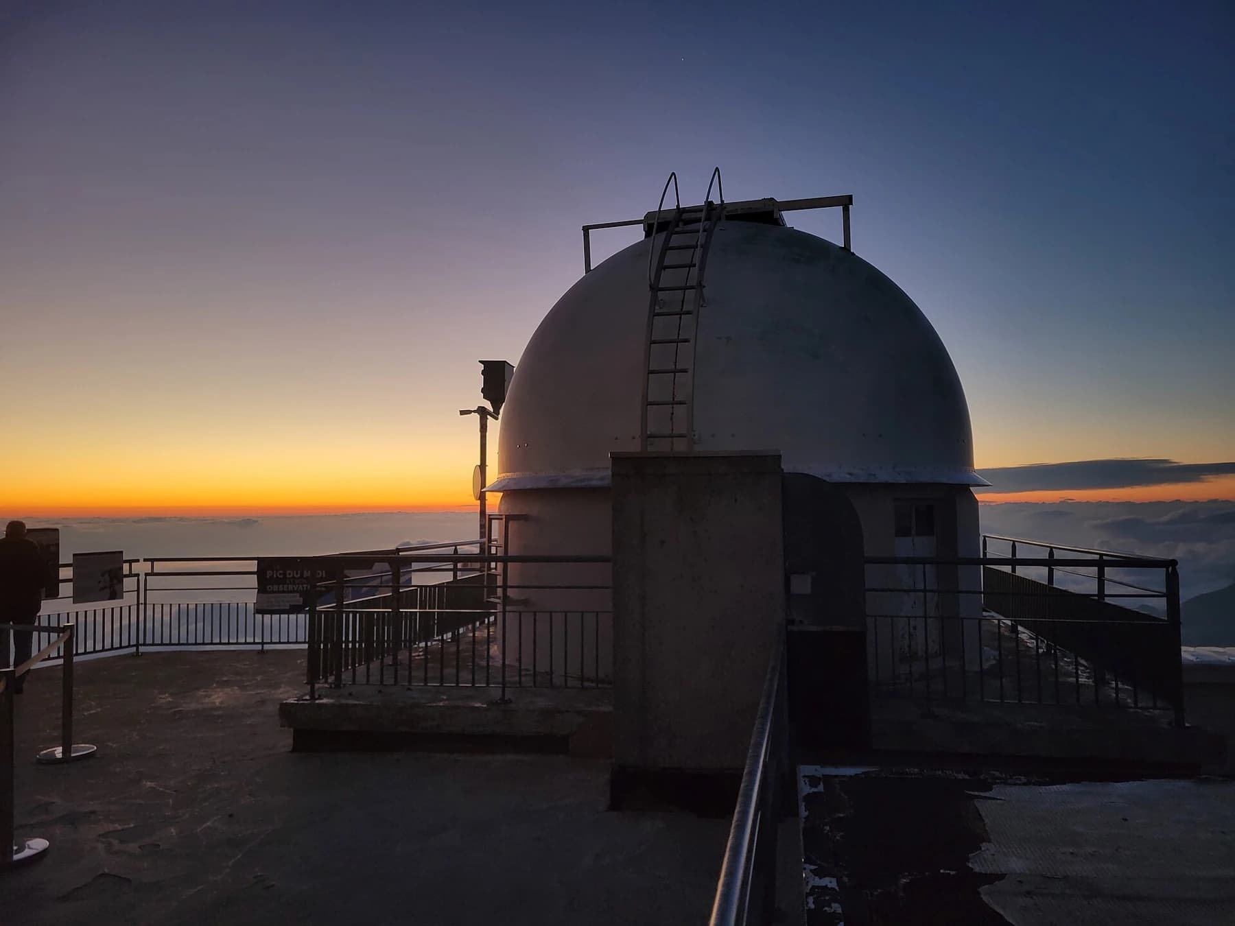 An observatory dome and terrace at Pic du Midi silhouetted against the orange horizon and blue sky after sunset.