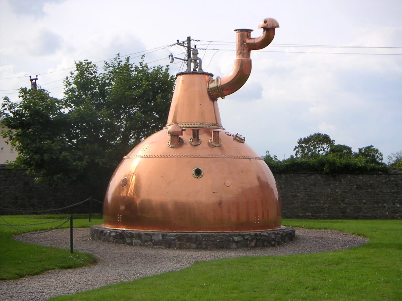 A large historical copper whiskey pot still displayed outdoors at the old Midleton distillery complex in County Cork, with trees behind it.