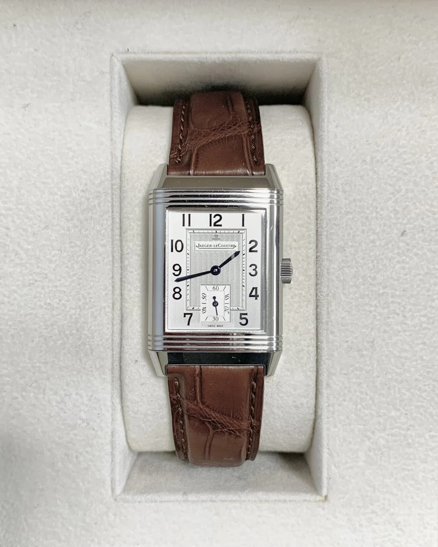 A rectangular Jaeger-LeCoultre Reverso with a silver-coloured Arabic-numeral dial and brown leather strap, resting in pale textured padding.