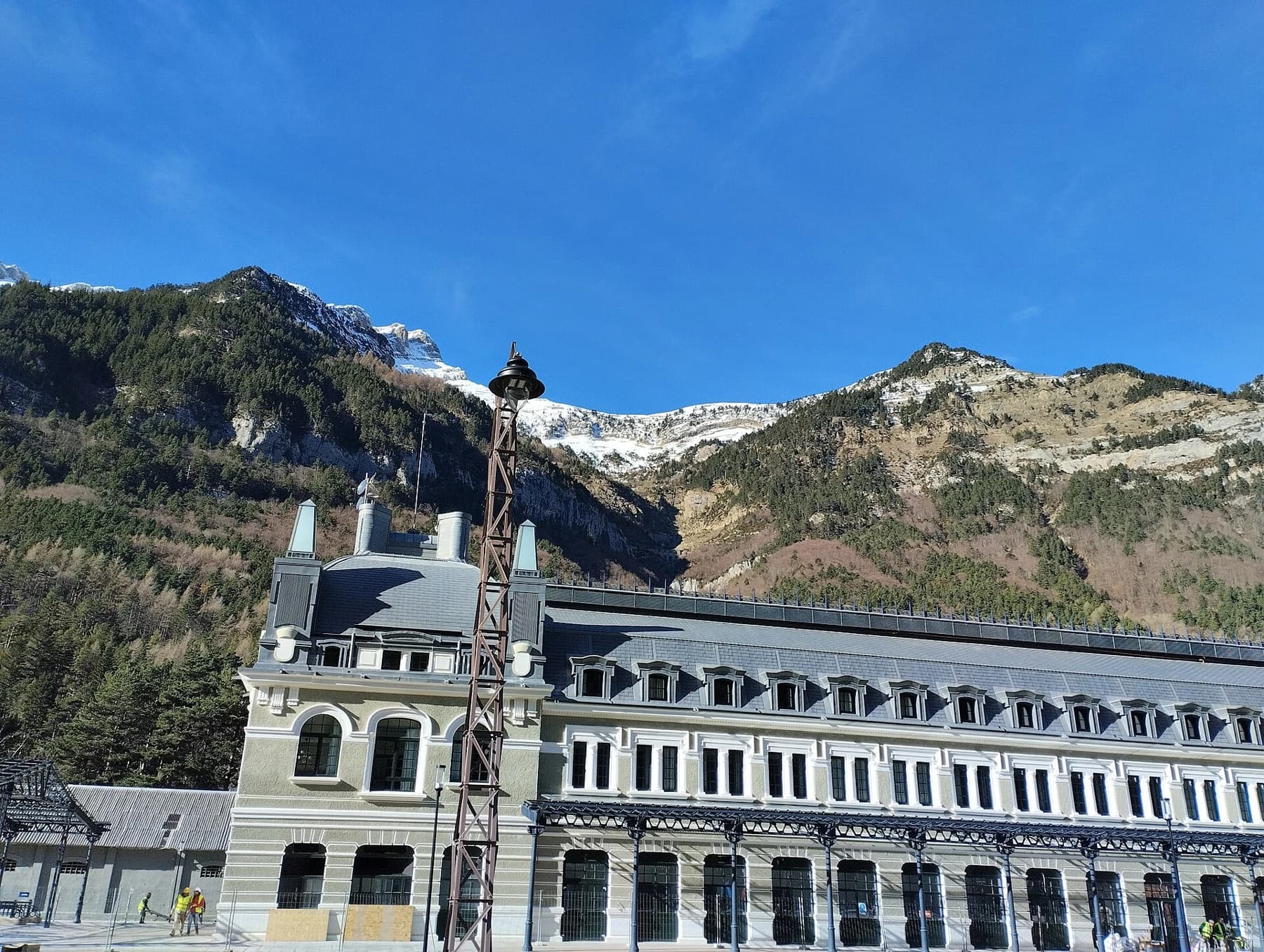 The central pavilion and long pale façade of Canfranc station beneath steep Pyrenean mountain slopes and a clear blue sky.