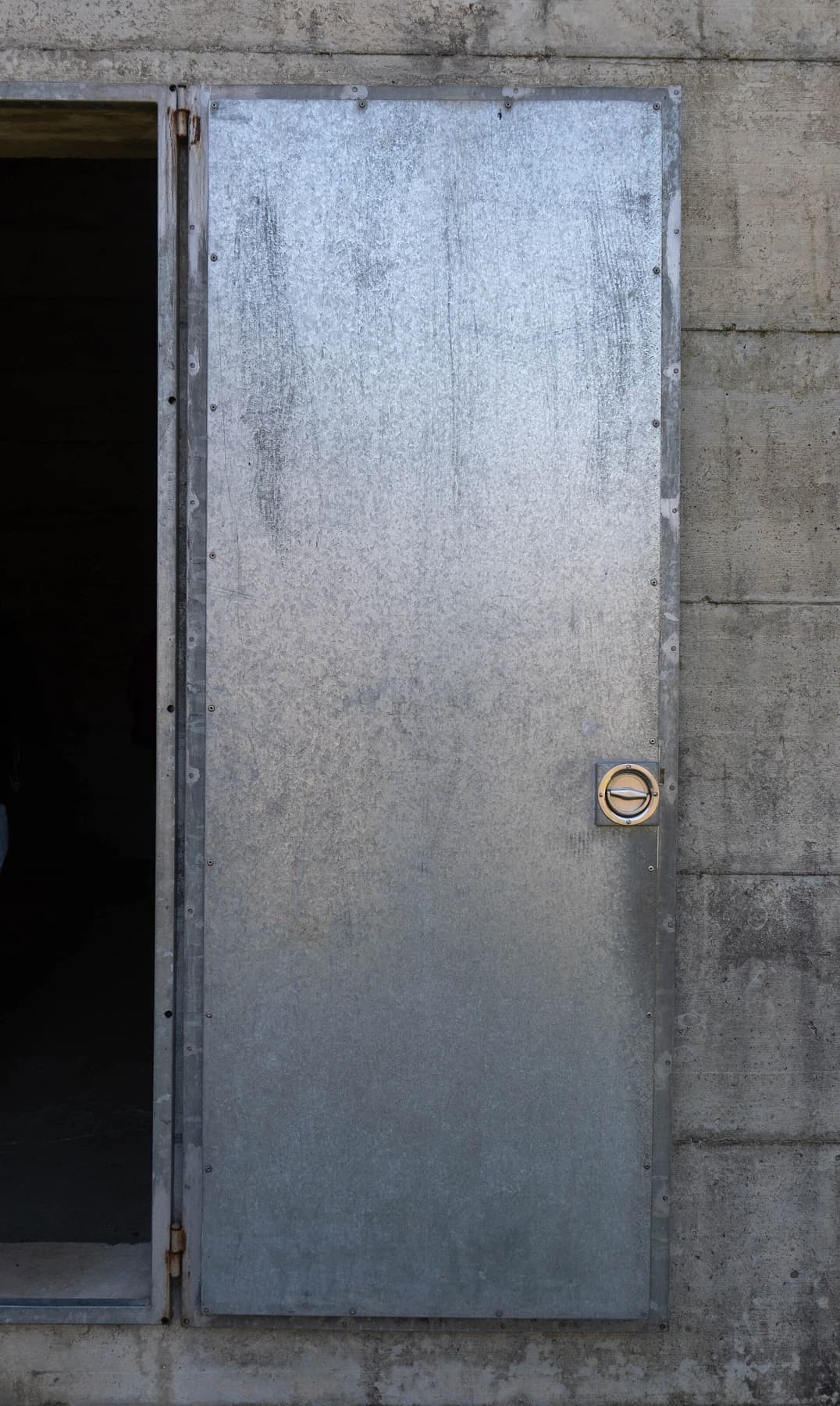 A plain metal entrance door stands open in the rough concrete wall of La Congiunta museum in Giornico, with a dark interior beyond.