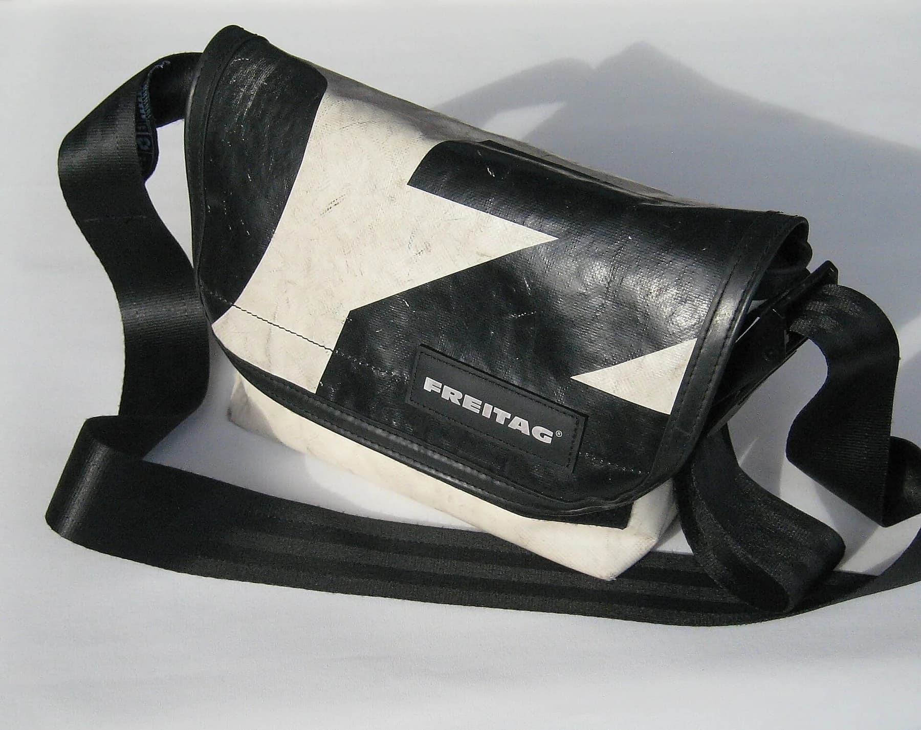 A black-and-white FREITAG messenger bag made from truck tarpaulin with a broad black strap, photographed against a pale background.