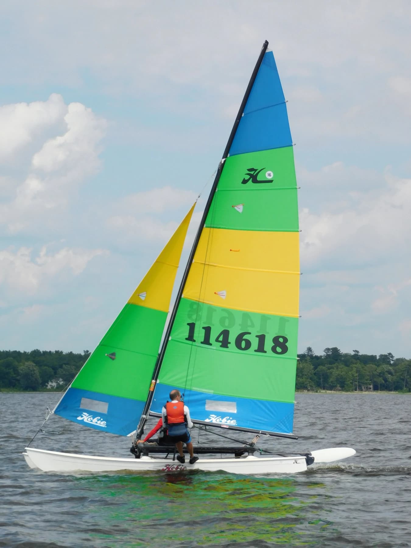 A Hobie 16 catamaran sailing with a blue, green and yellow sail, two white hulls and a sailor visible above the water.