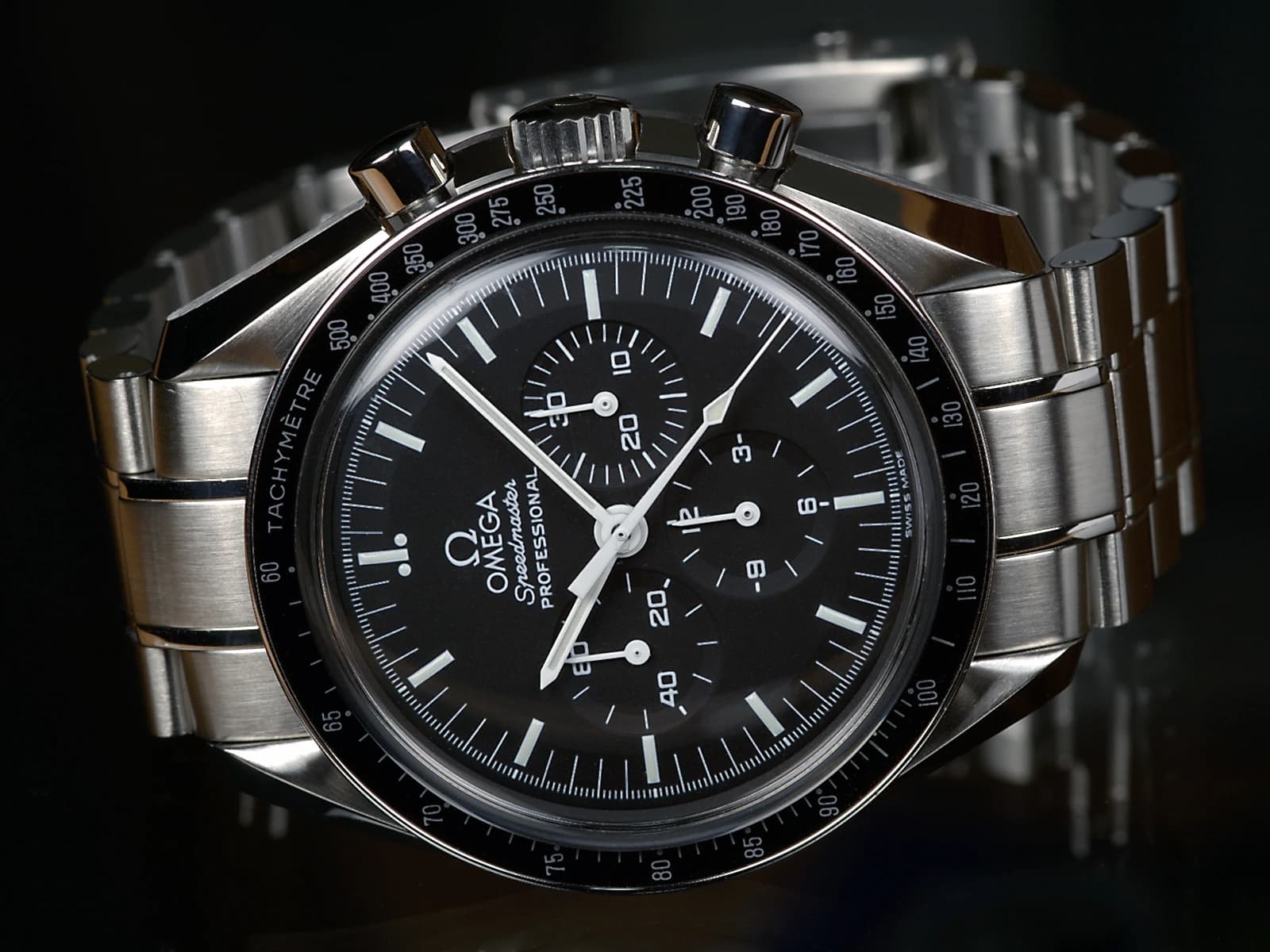An Omega Speedmaster Professional chronograph with a black dial, three subsidiary dials, tachymeter bezel and steel bracelet against a dark background.