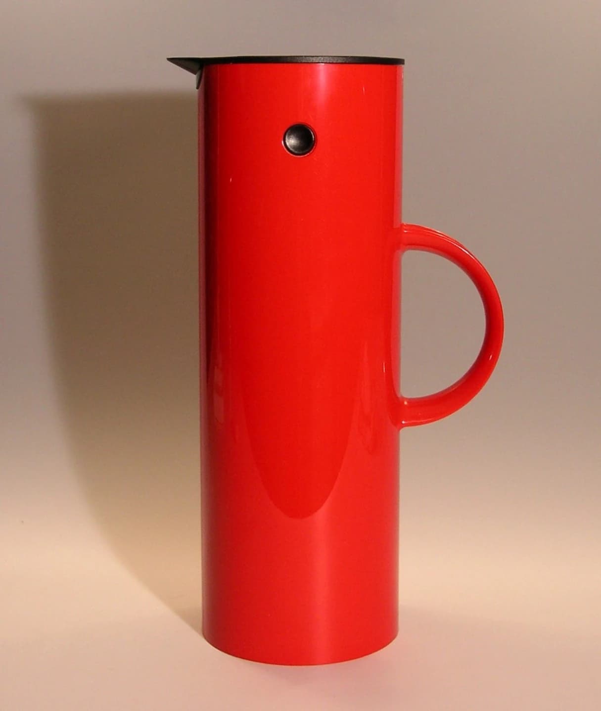 A red Stelton vacuum jug designed by Erik Magnussen, with a black tipping stopper, a circular side handle and a tall cylindrical body.