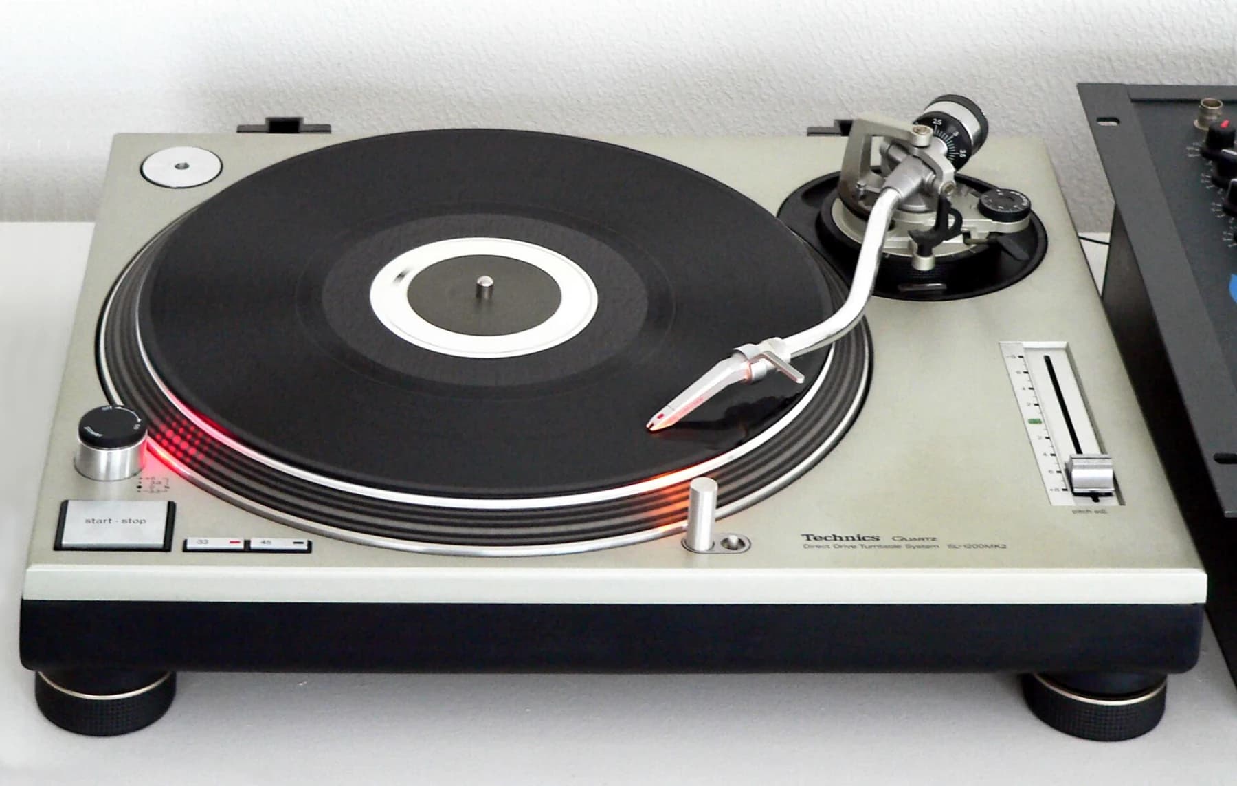 A silver Technics SL-1200MK2 turntable with a record, curved tonearm, pitch fader and red strobe light visible against a pale background.